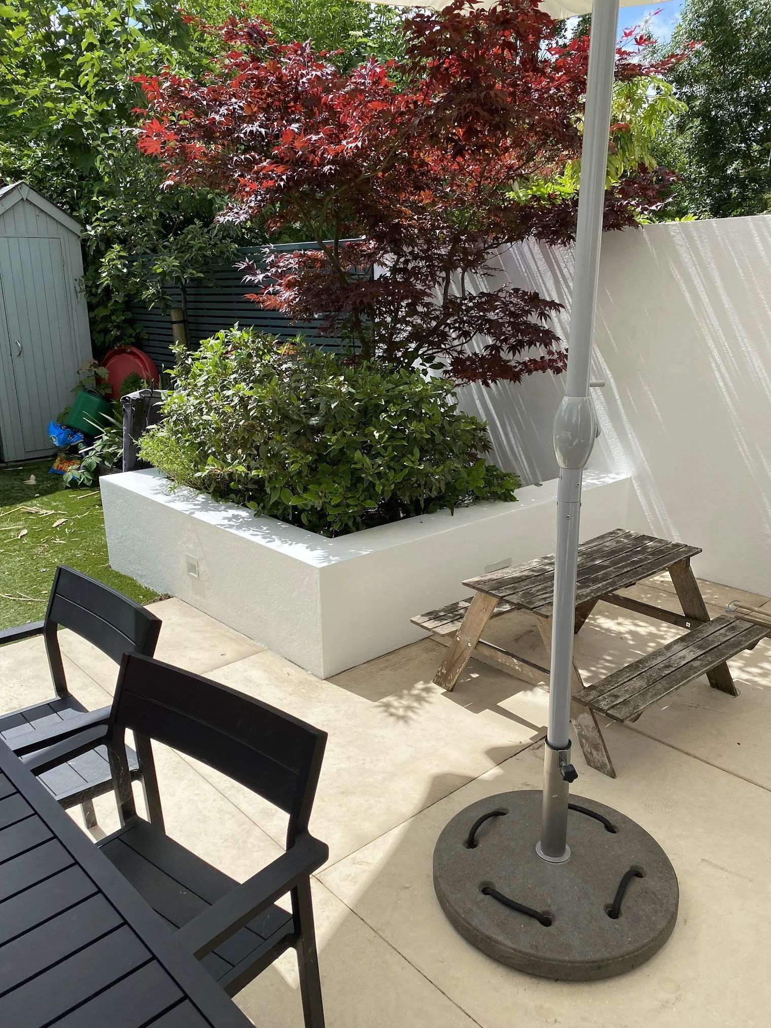 A patio scene with black dining chairs, a wooden bench, a large beige patio umbrella, a white raised garden bed with a red-leaved Japanese maple tree and green shrubbery, a white privacy fence, a small gray shed, and gardening supplies in the backgro
