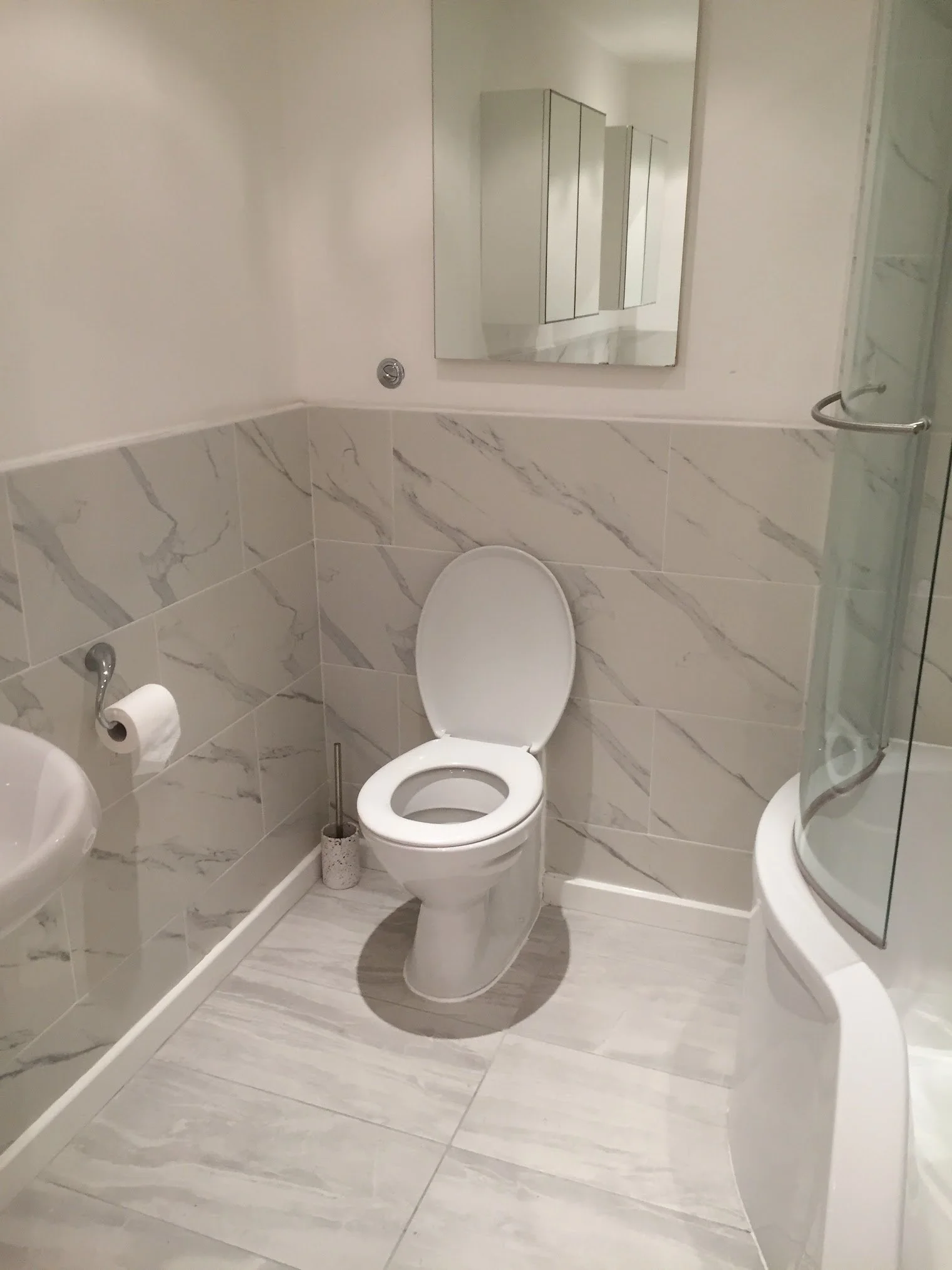 A small bathroom with a toilet, a mirror, a shower with glass enclosure, and marble-tile walls and floor.