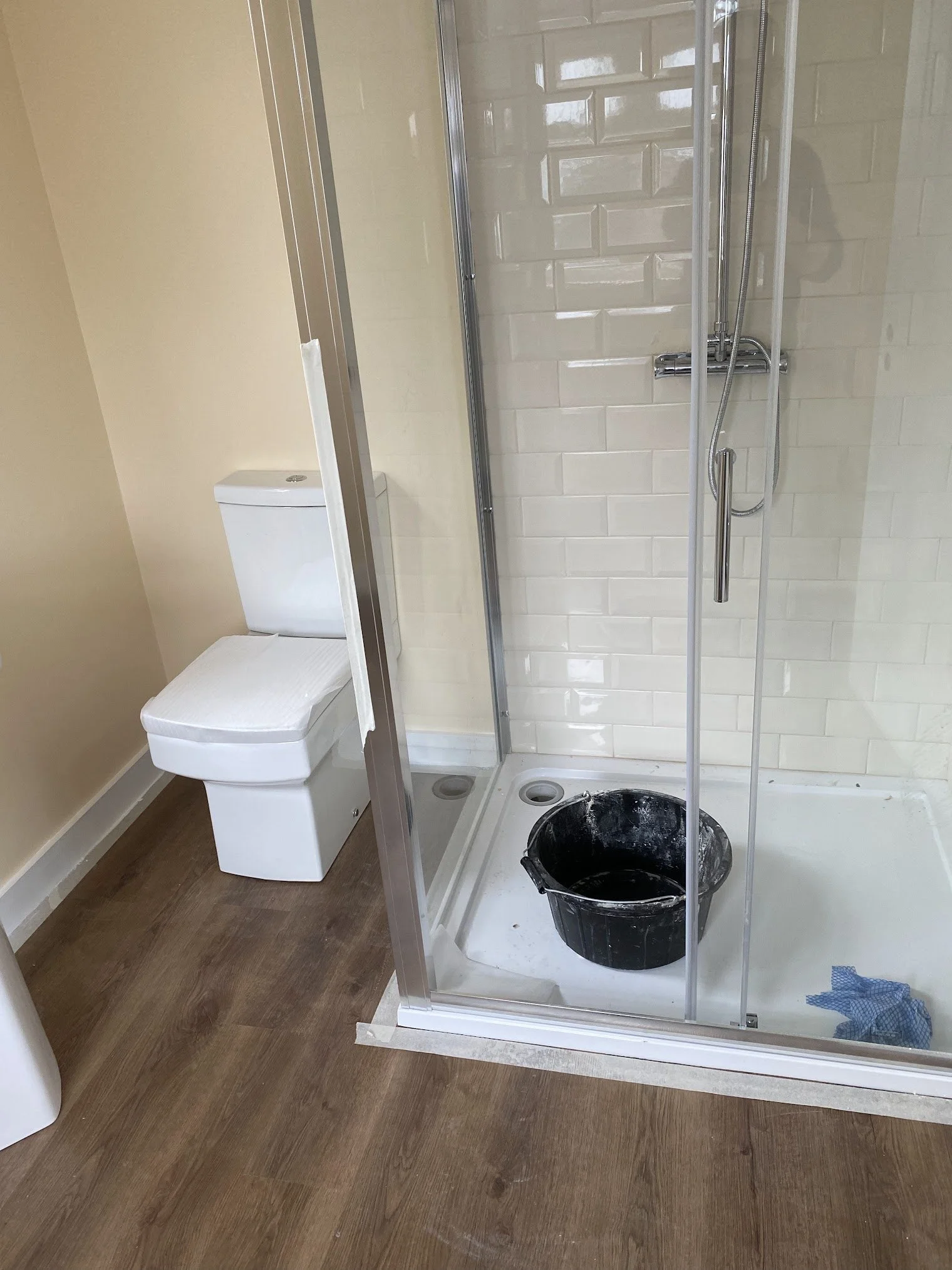 A bathroom with a toilet, a partially installed shower enclosure, and a bucket filled with black paint or liquid, along with some cloths on the floor.