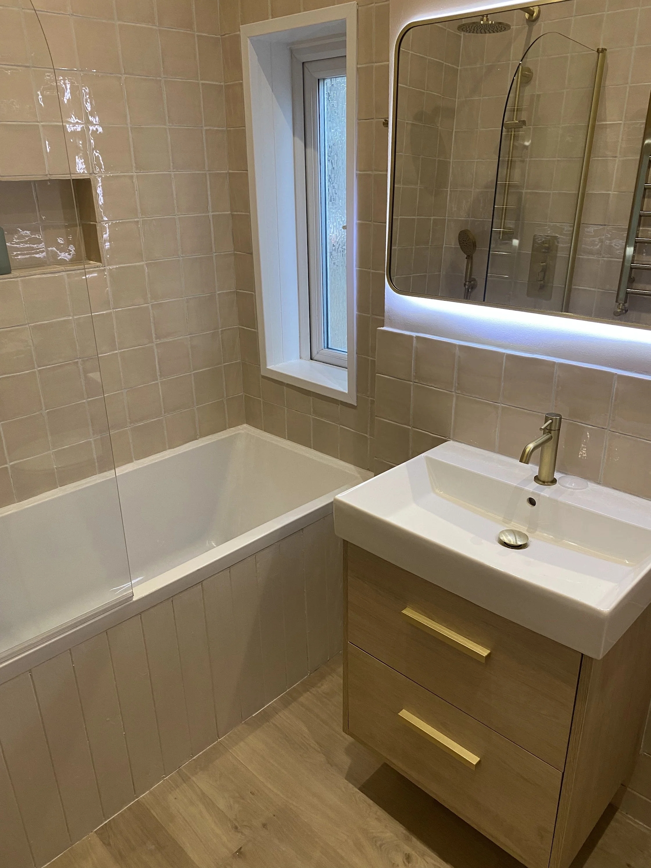 Bathroom with beige tiled walls, a small rectangular sink with a gold-colored faucet and drawer, a mirror with LED lighting, a window, a bathtub, and a shower with a rainfall head and handheld shower.