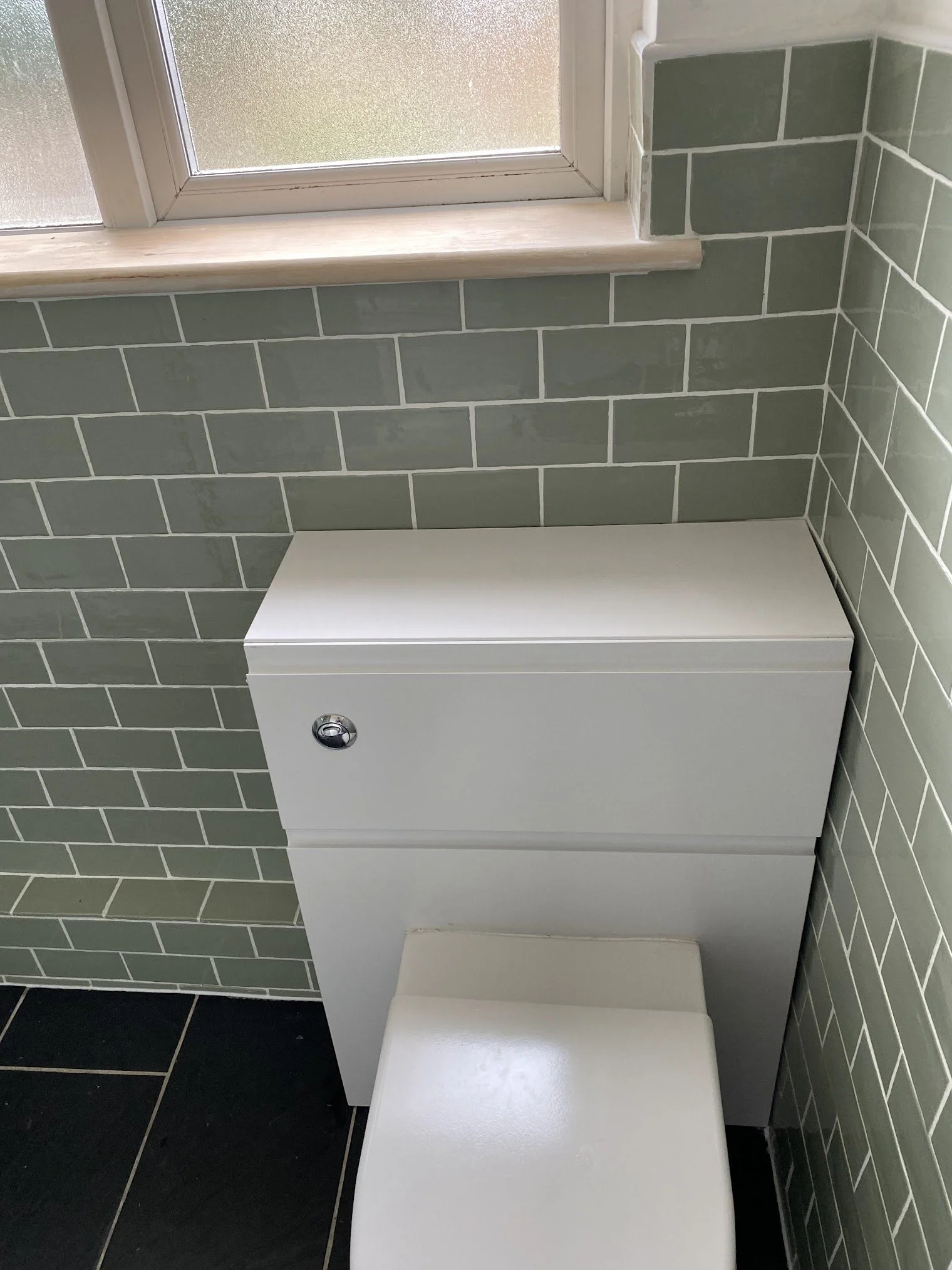 A white modern toilet with a flush button on a flush plate, positioned against a wall with green subway tiles, and adjacent to a frosted window with a wooden windowsill.
