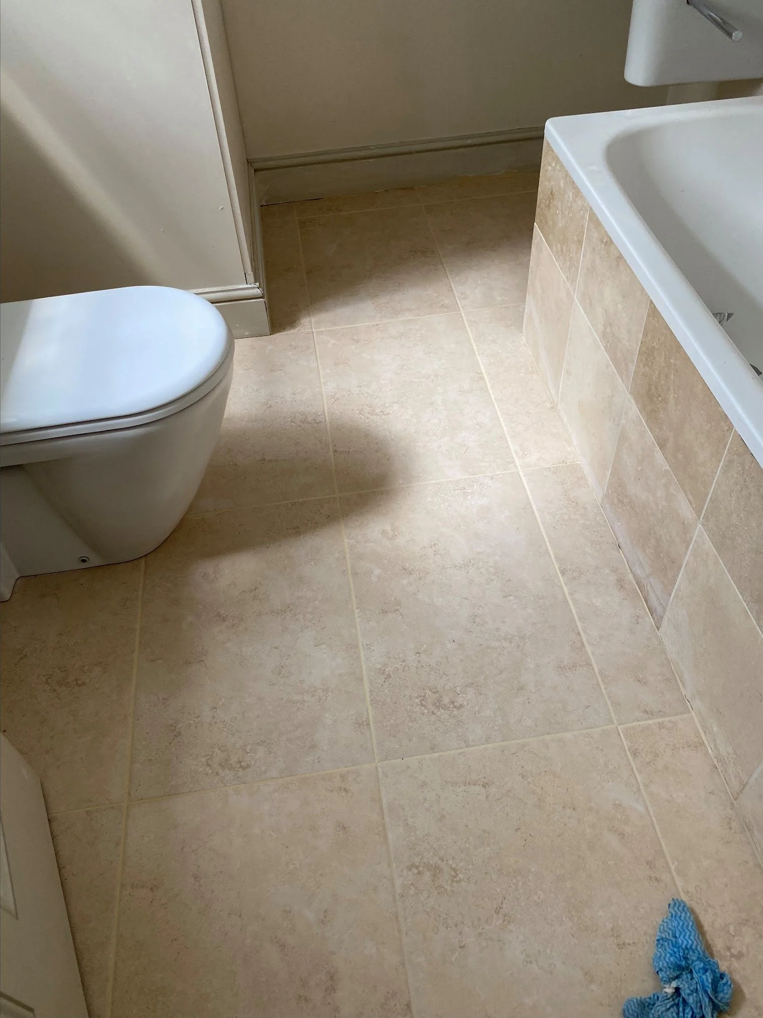 A bathroom with beige tiled floor, a white toilet in the lower left corner, a toilet paper holder on the wall to the right, and a crumpled blue cloth on the floor in the lower right corner.