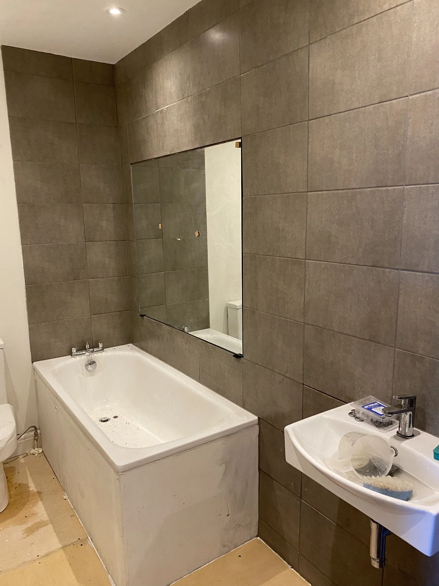 A partially constructed or renovated bathroom with brown tiled walls, a white bathtub, a small white sink with a mirror above, and various construction materials and tools scattered around.