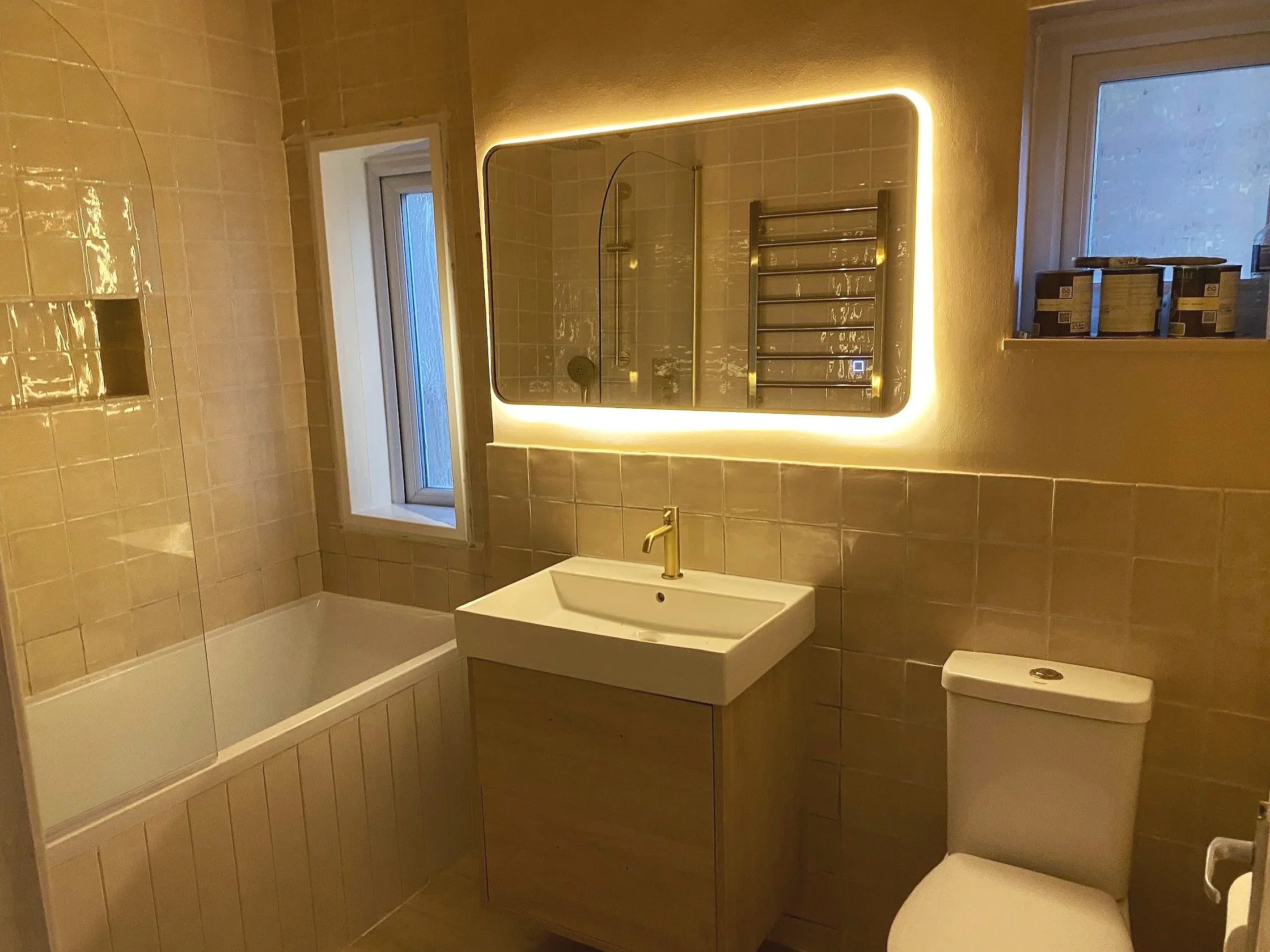 A modern bathroom with beige tiled walls, a heated towel rack, a rectangular mirror with backlighting, a window, a white sink with a gold faucet, a toilet, and shelves with boxes.