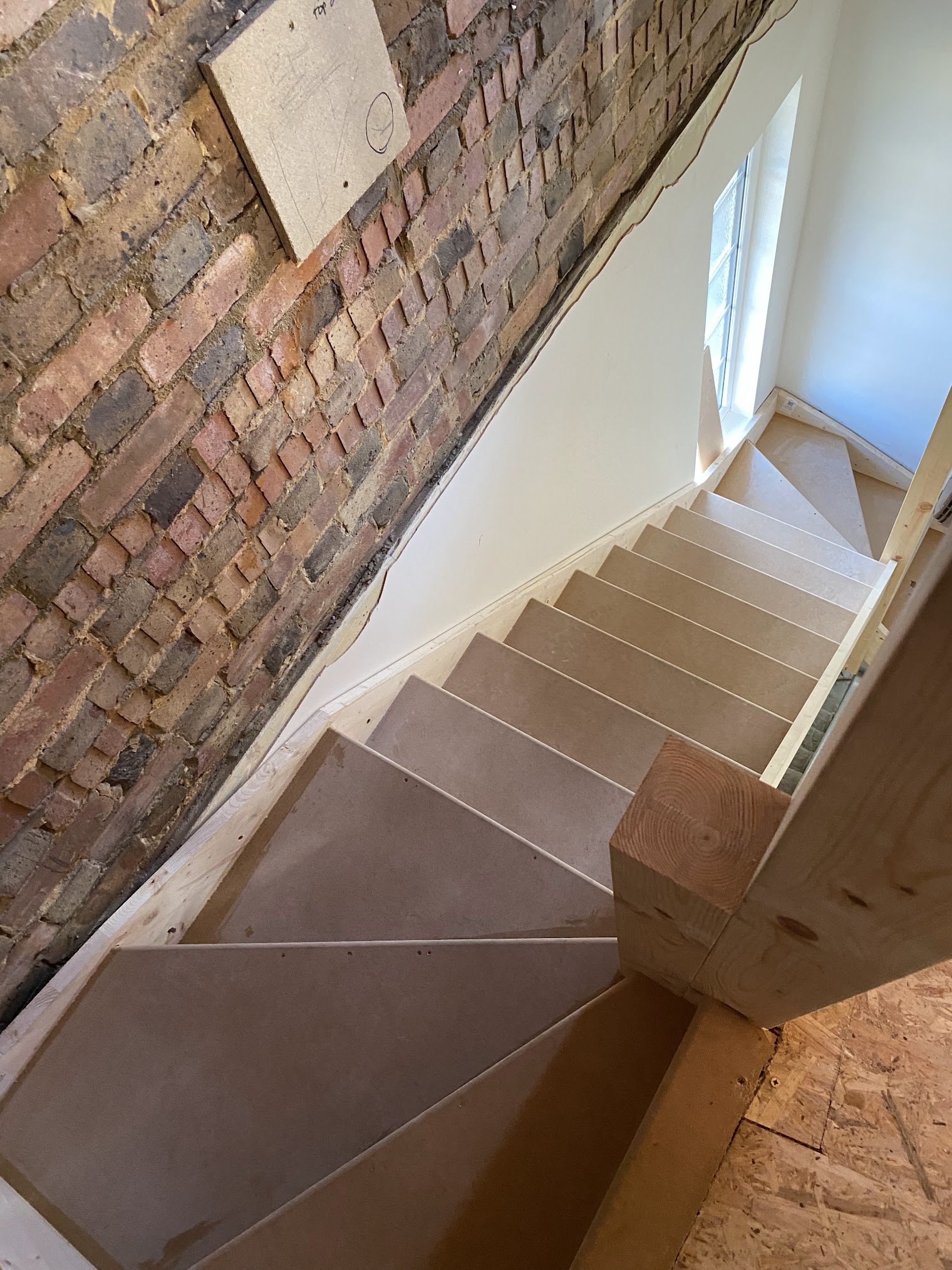 View of a staircase with unfinished wooden steps, a brick wall on the left, and a window allowing natural light to enter the space. Construction in progress.