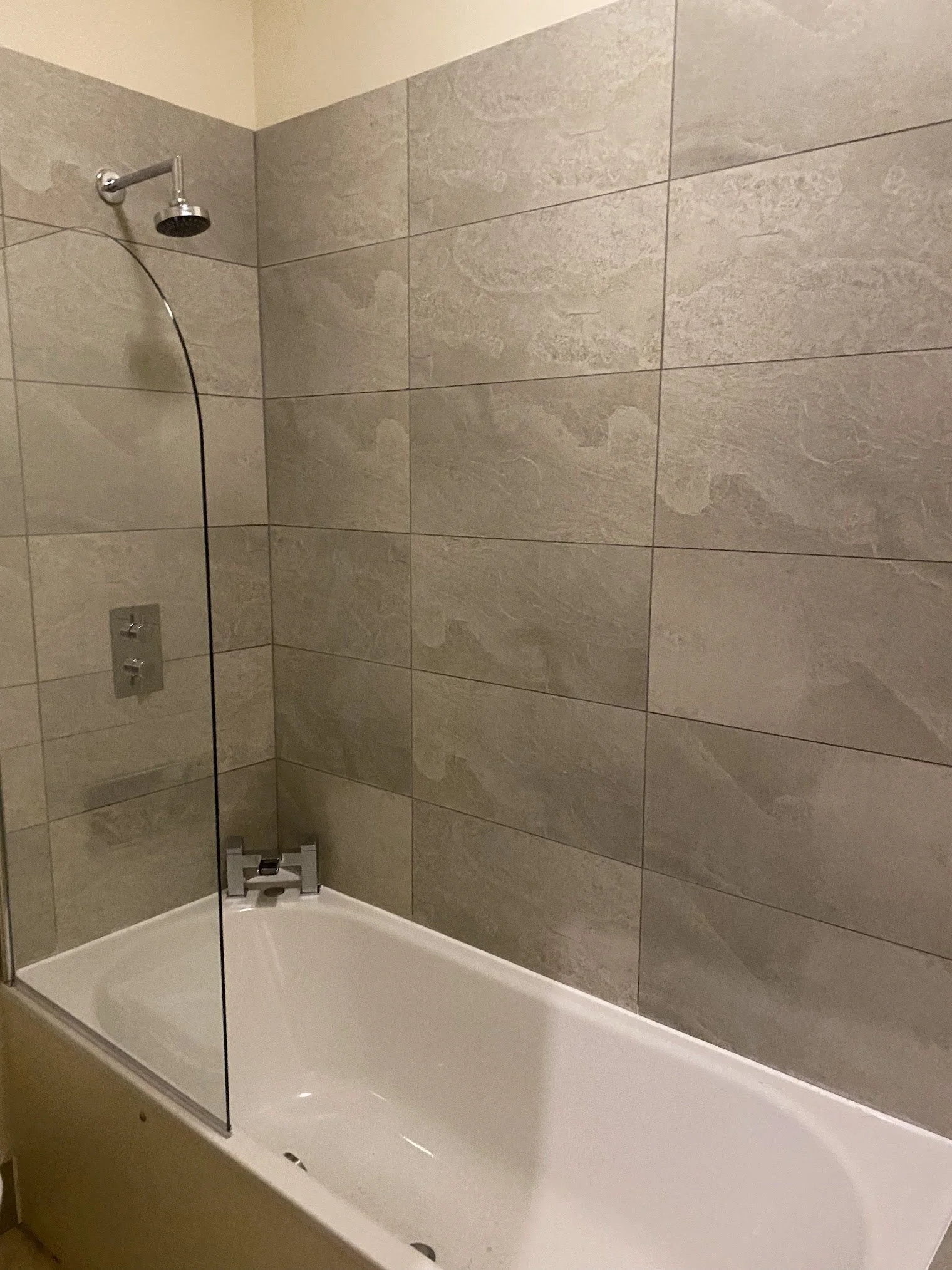 A bathroom shower with gray tiled walls, a chrome showerhead, a glass door, and a white bathtub.