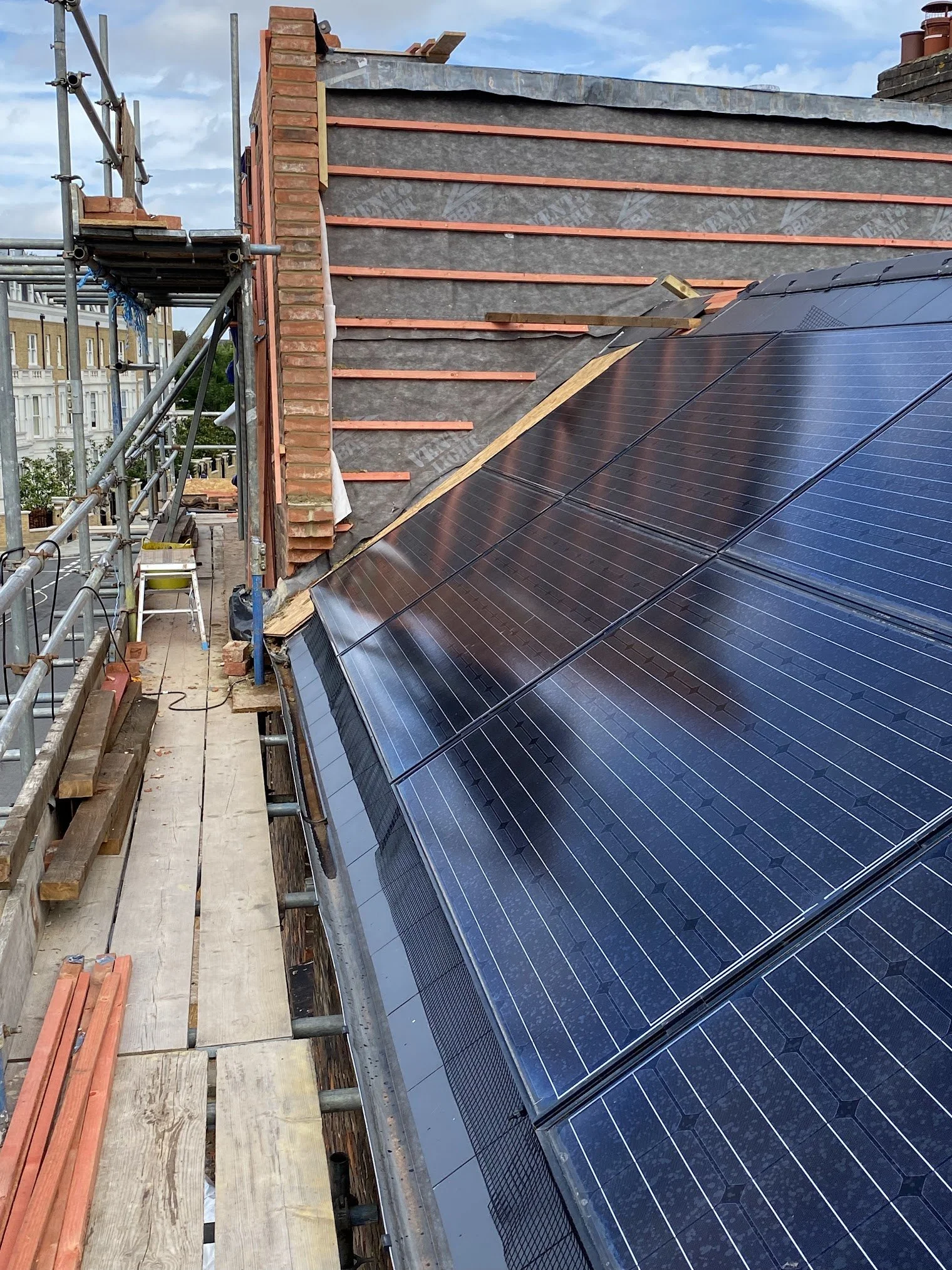 A rooftop with solar panels installed, part of the roof showing bricks and roofing materials, scaffolding and construction tools present, with a partly cloudy sky in the background.