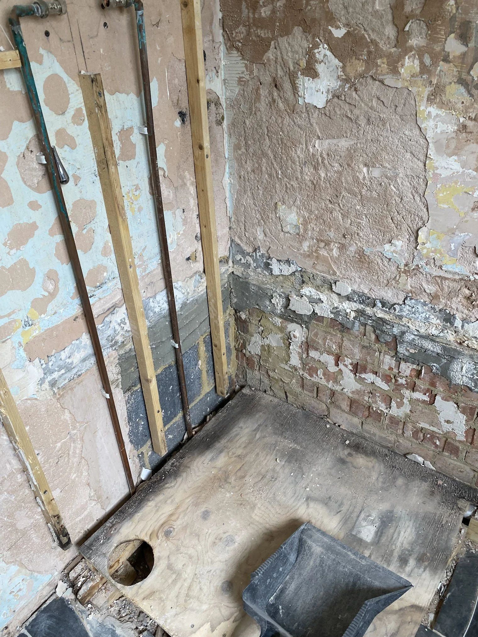 Interior of a room under renovation with exposed brick walls, plumbing pipes, a cutout in a countertop, and construction tools.