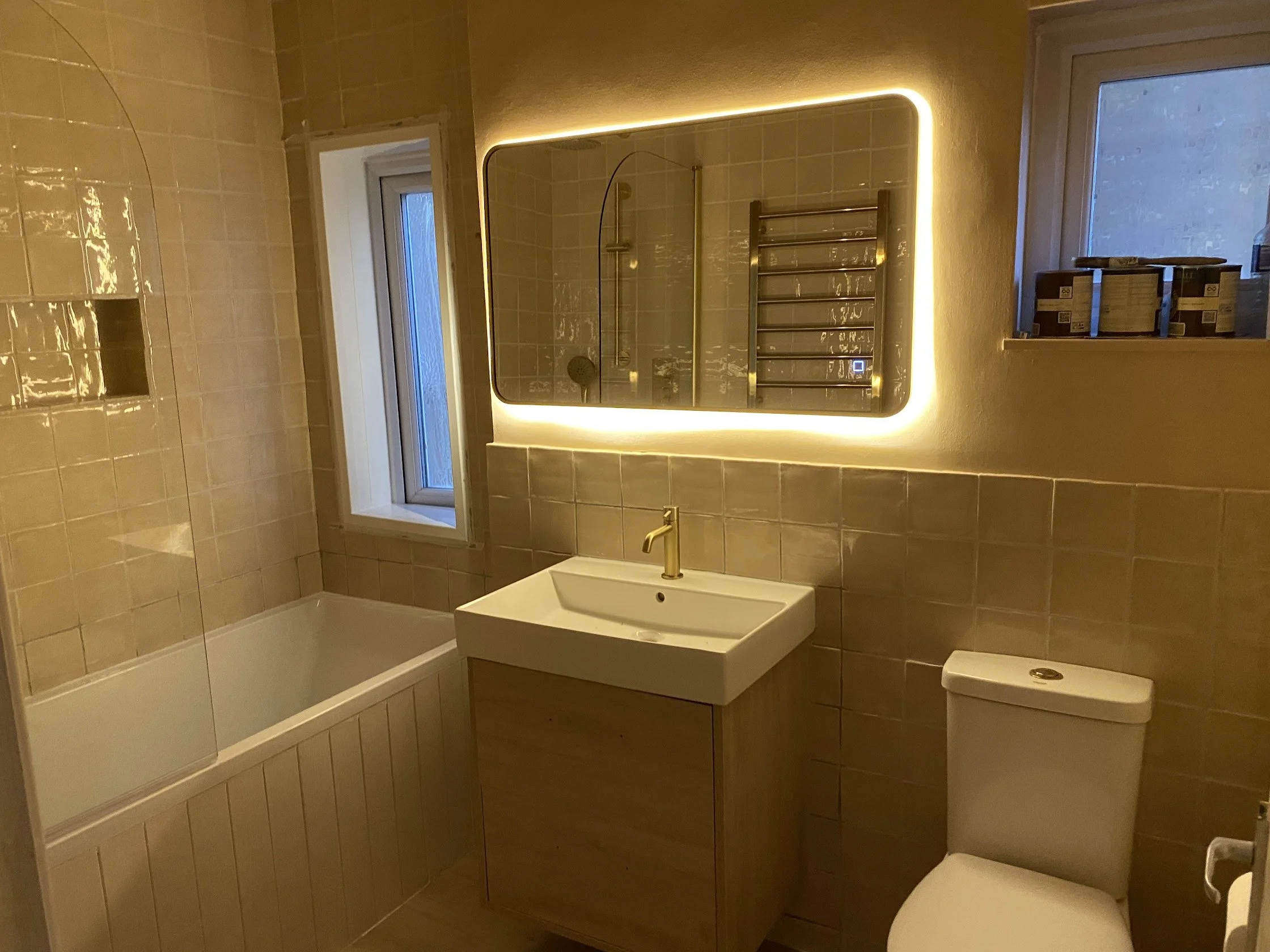 A modern bathroom with a white bathtub, a wall-mounted sink with a gold faucet, a toilet, and a large mirror with LED backlighting. There is a small window above the bathtub, and a shelf with some boxes on it.