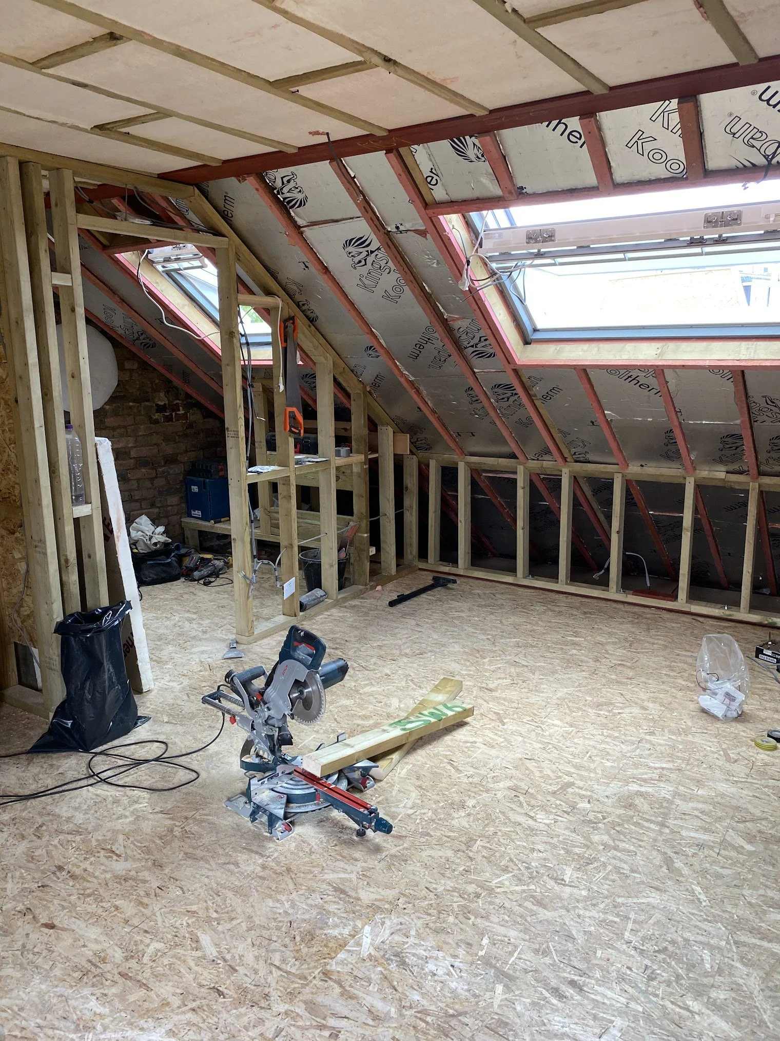 Attic or attic renovation space with unfinished wood framing, skylight windows, construction tools, debris, and insulation panels.