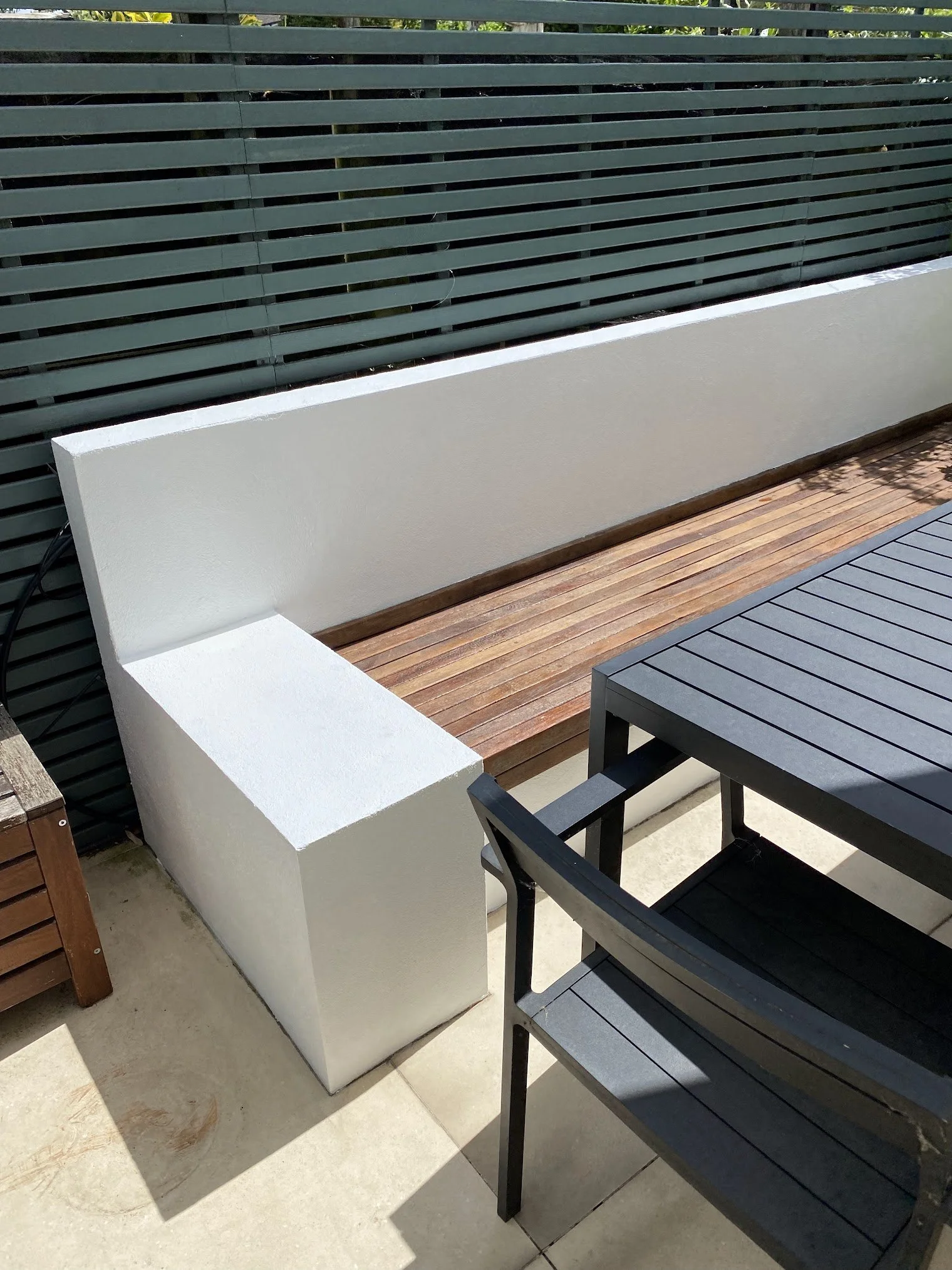 A modern outdoor balcony with a wooden floor, white wall, and black furniture, including a table and chairs, overlooking a green garden.
