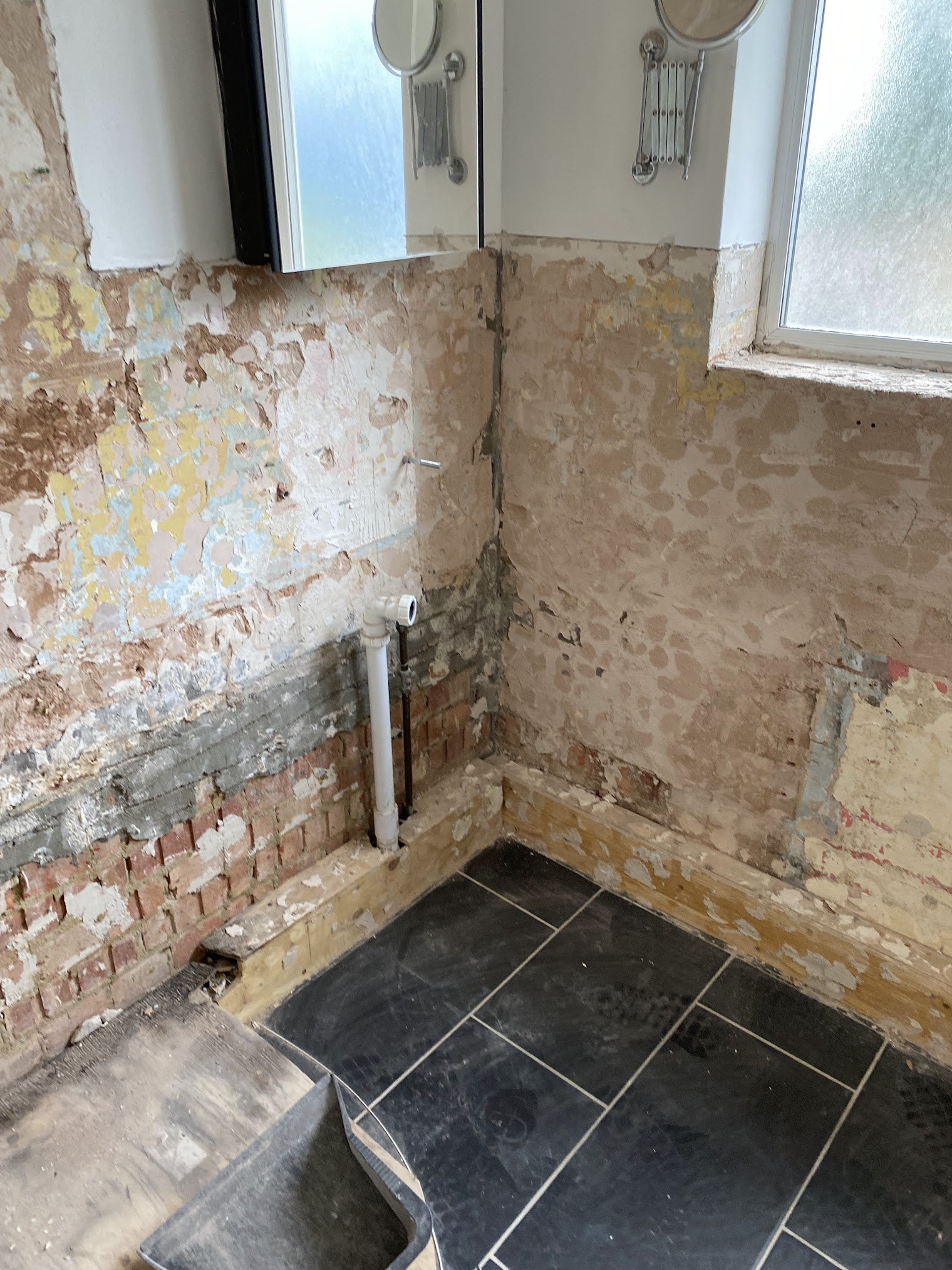 A bathroom under renovation showing exposed brick and plaster walls, a black tiled floor, and plumbing pipes. A mirror cabinet and frosted window are visible at the top.