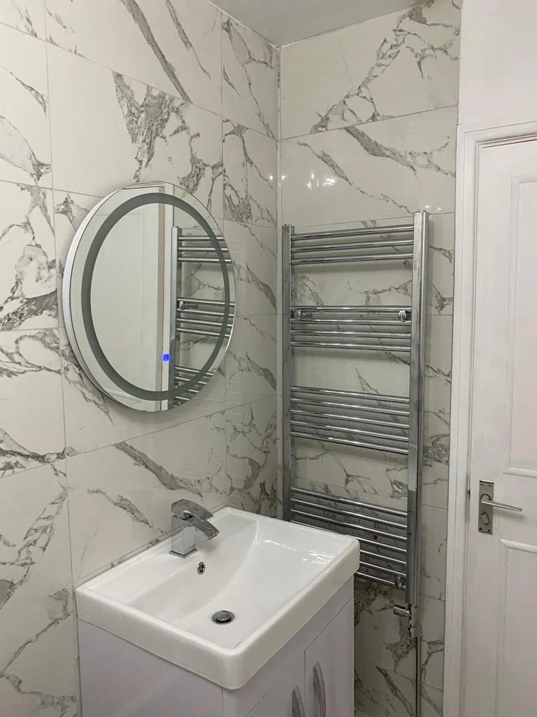 A bathroom with marble-patterned tiles, a mirror, a white sink with a faucet, and a chrome heated towel rack.