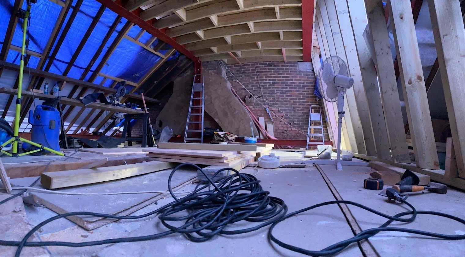 Interior view of an attic under construction with exposed wooden beams, tools, and building materials scattered around.