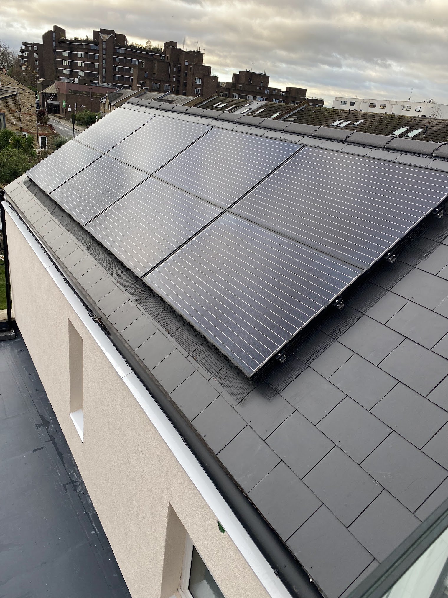 View of a rooftop with installed solar panels, apartment buildings in the background, and cloudy sky.