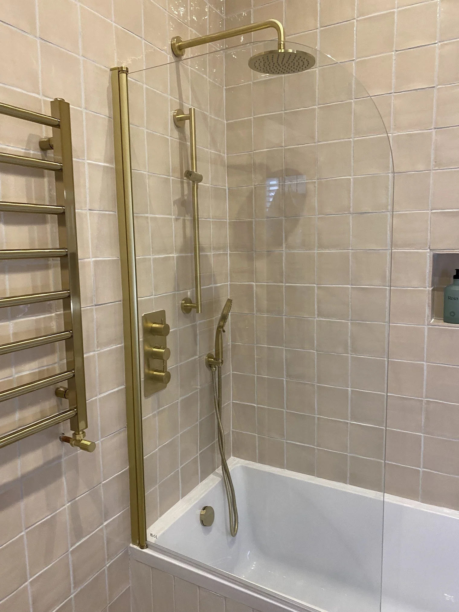A beige tiled shower with a brass showerhead, a brass handrail, and multiple brass controls, including a handheld shower wand, in a bathroom.