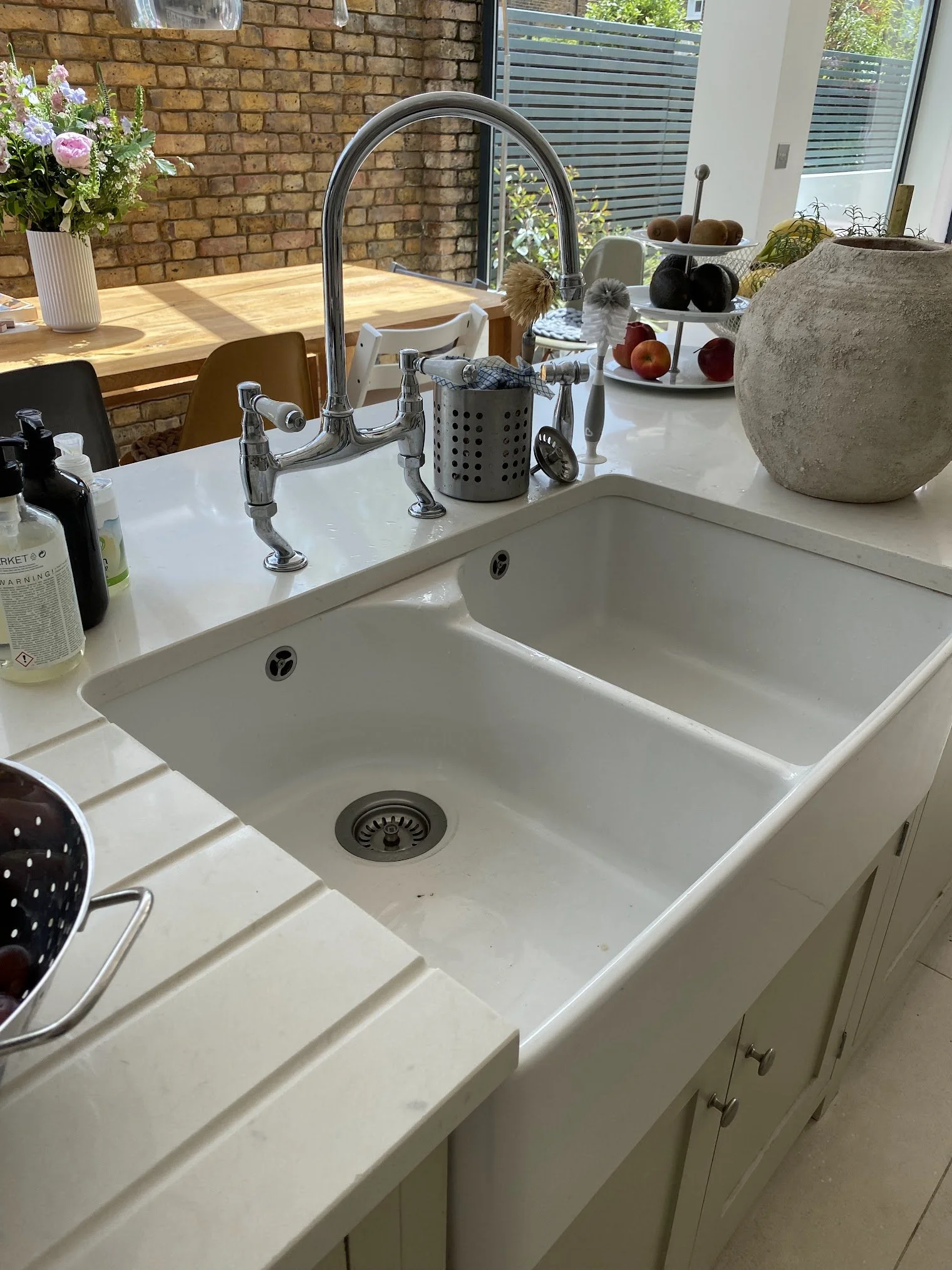Kitchen sink with soap dispensers, brush, and a large stone pot on the counter, with a tiered tray of apples and avocados, and a potted plant in a white vase nearby, with a brick wall and large window in the background.