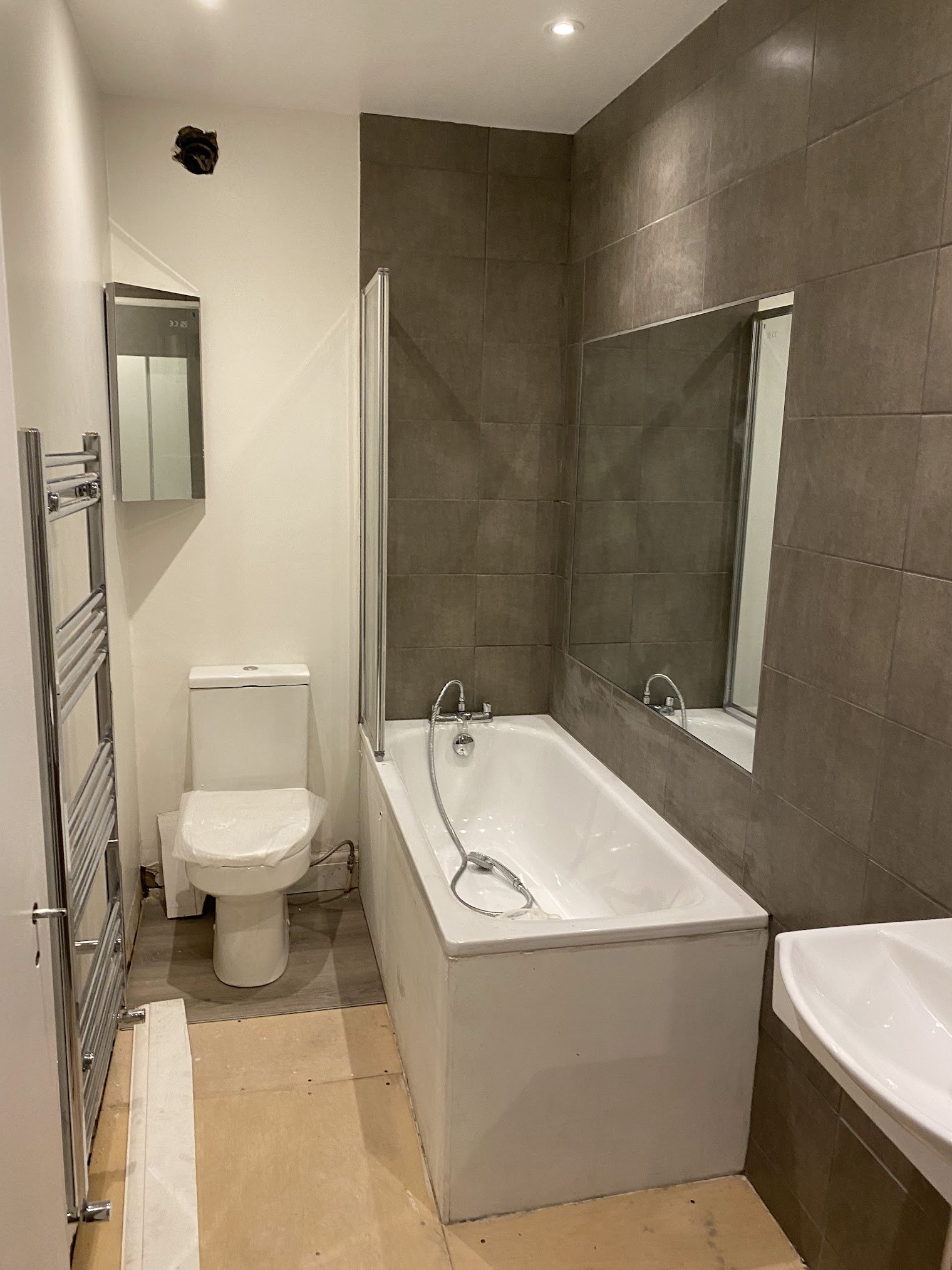 Bathroom with a white bathtub, gray tiled wall, toilet, small mirror, and towel warmer in a modern design.