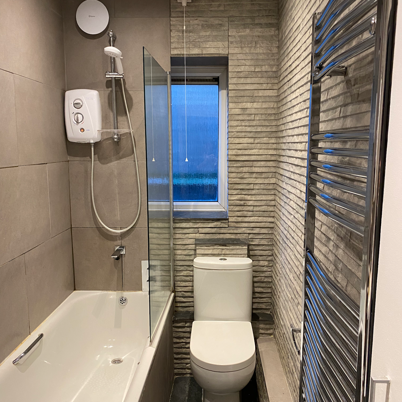 A compact bathroom with a bathtub and shower, a toilet, a window with frosted glass, a heated towel rack, and decorative brick-patterned wall tiles.