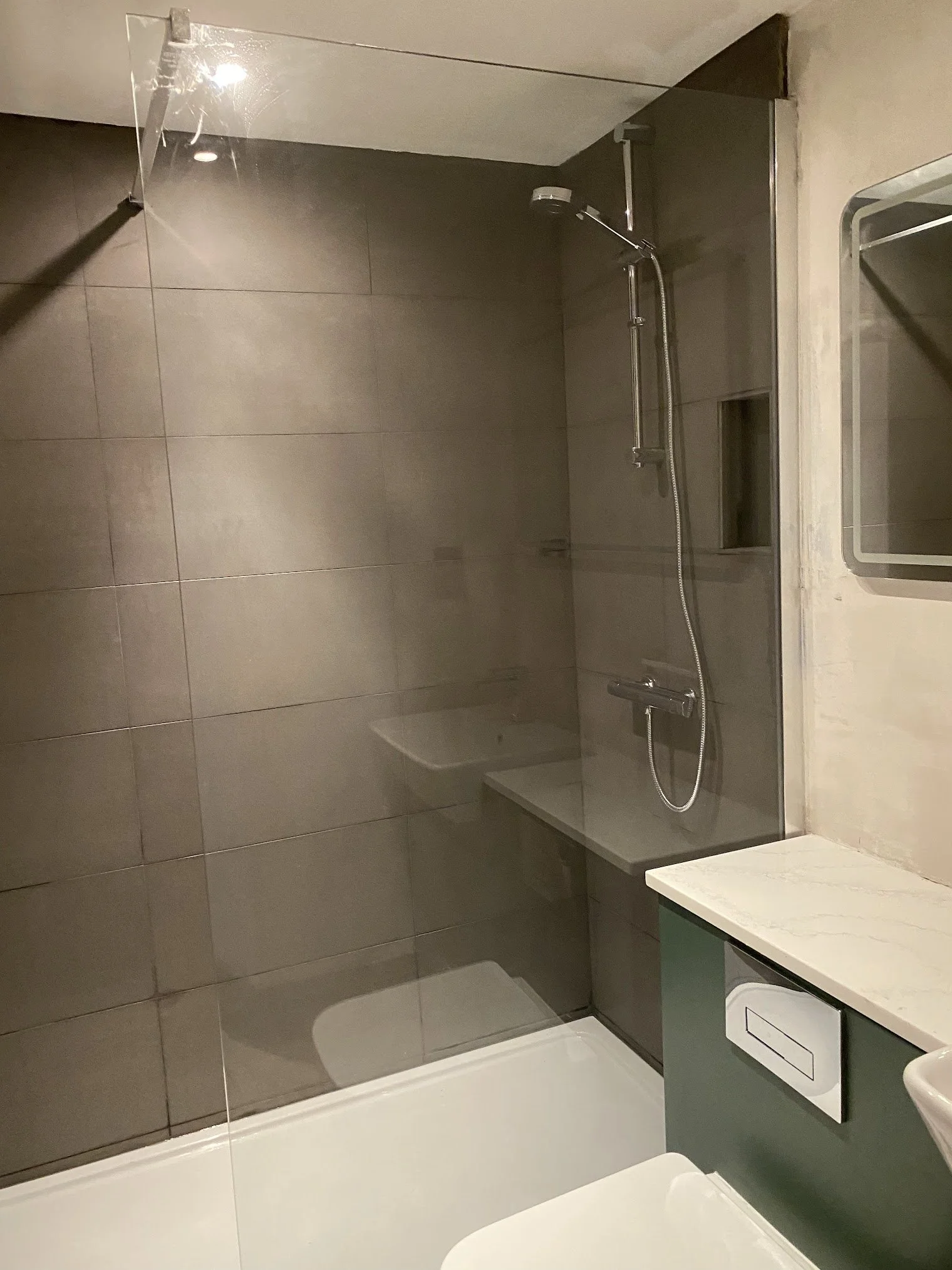 Modern bathroom with gray tiled walls, a shower area with a glass partition, a mounted showerhead, a mirror, a white sink, and a washing machine.