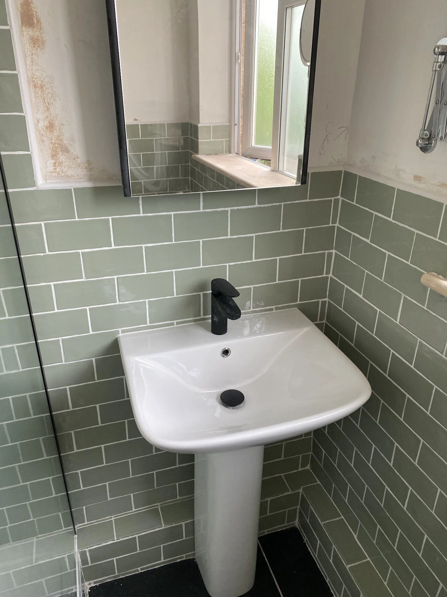 A bathroom with green subway tiles on the walls, a white pedestal sink with a black faucet, a mirror, and a window. The walls show some unfinished areas and markings.
