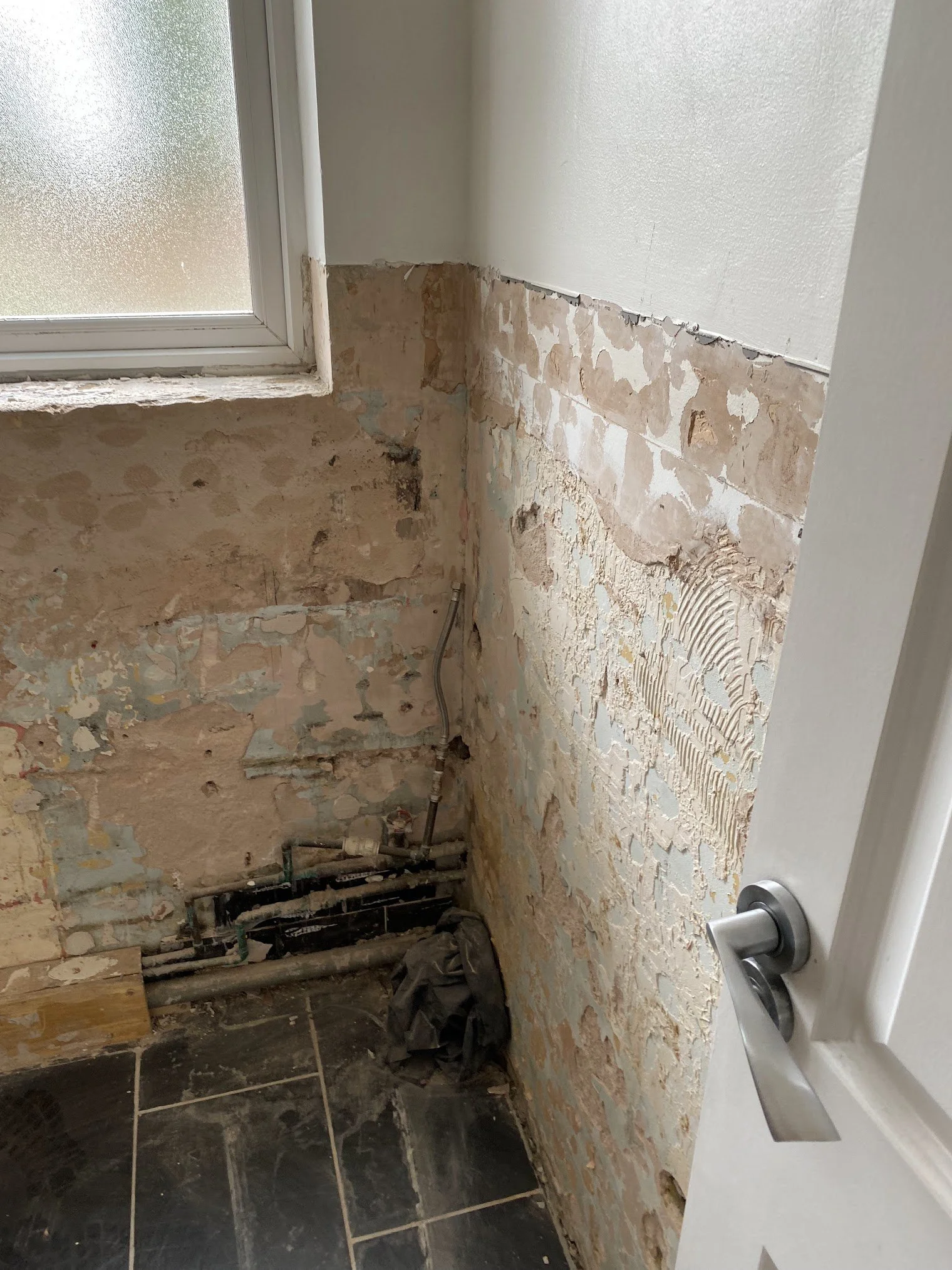 A partially gutted wall in a bathroom, showing exposed brick and plumbing pipes, next to a frosted glass window and a white door with a silver door handle.