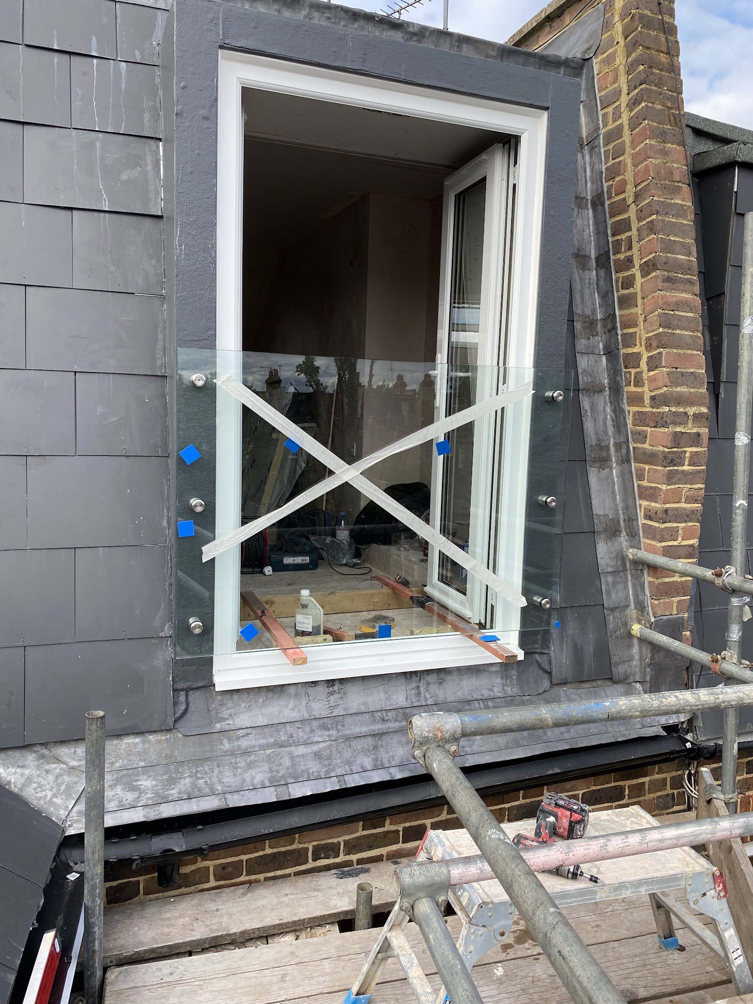 A construction site showing a building with mostly gray and brick exterior walls. A large window is being installed with visible scaffolding and tools around. The window frame is white, and the construction appears to be in progress with safety tape 