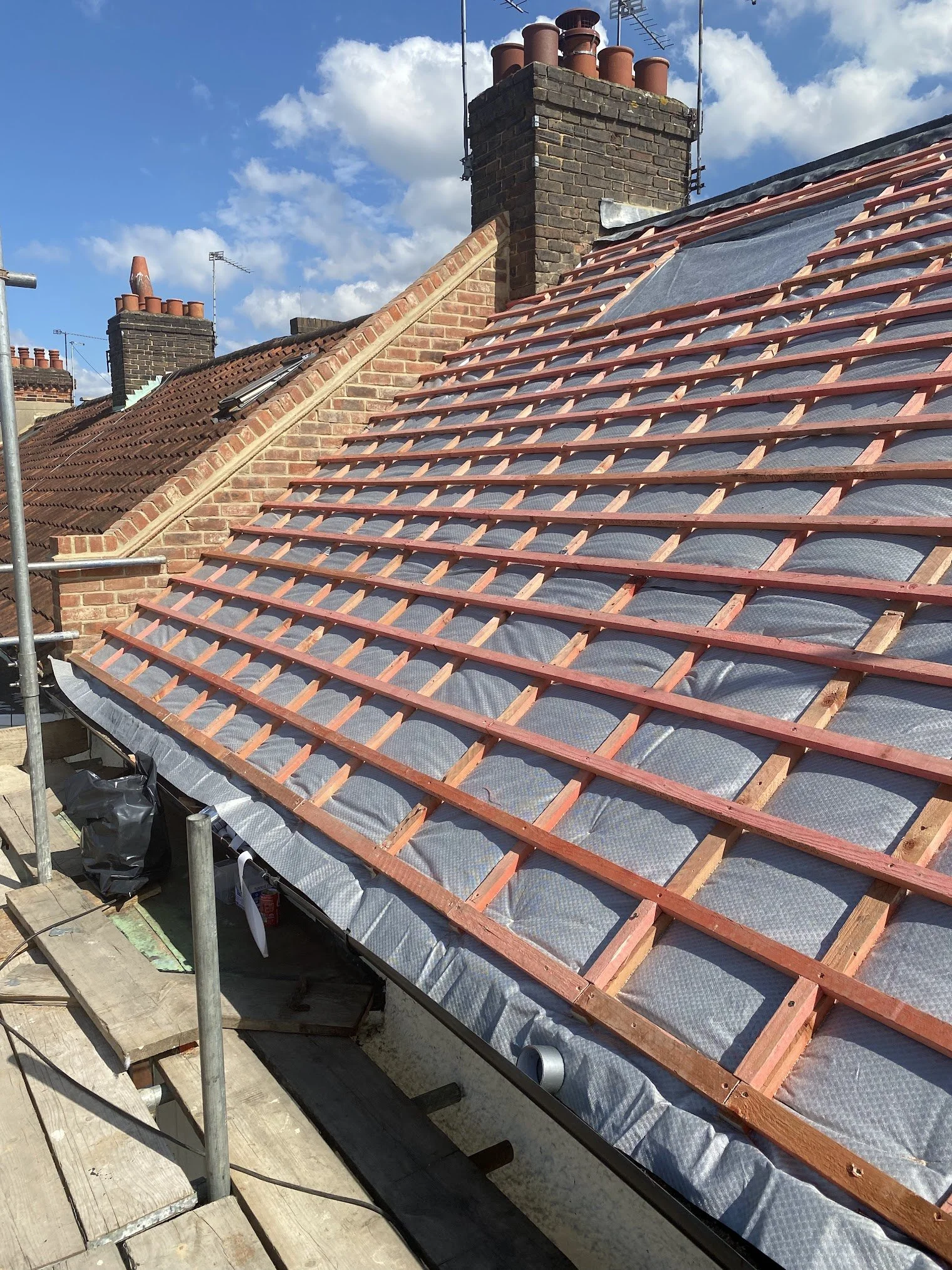 Roof under construction showing wooden battens installed over insulation, with brick chimneys and a partially covered section, under a partly cloudy sky.
