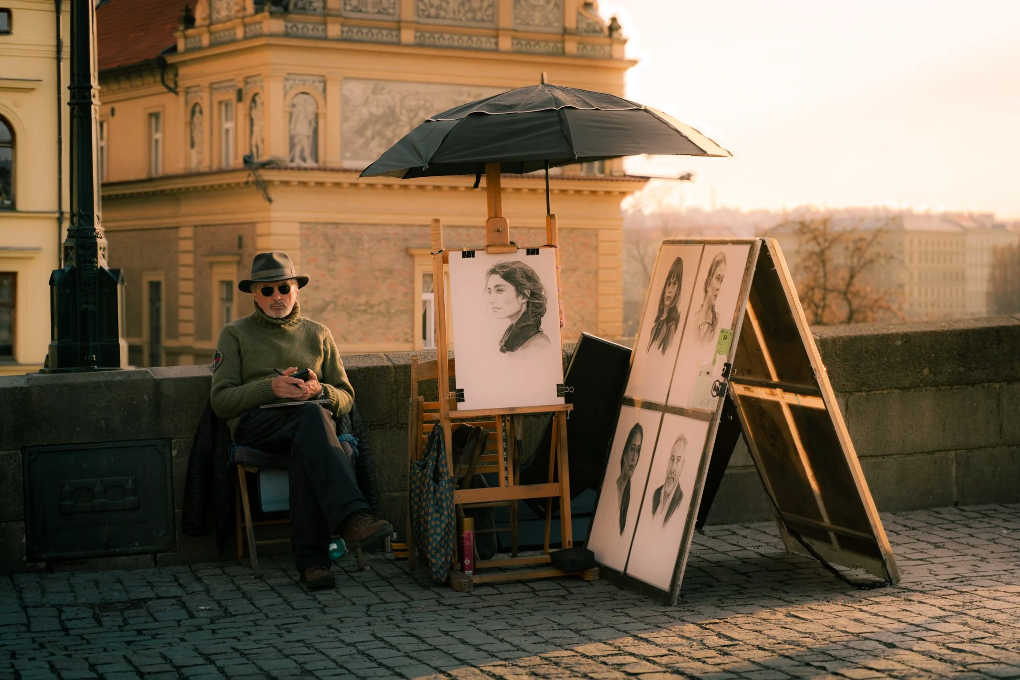 An elderly man with sunglasses and a hat sitting on a bench next to street art sketches outside in golden sunlight. Artist’s drawings of women are displayed on easels and boards next to him, with a historic building in the background.