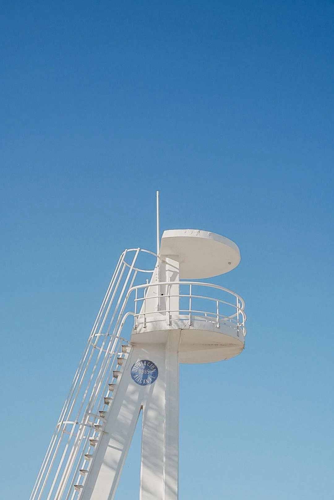 A white observation tower against a clear blue sky with a small circular platform and railing.