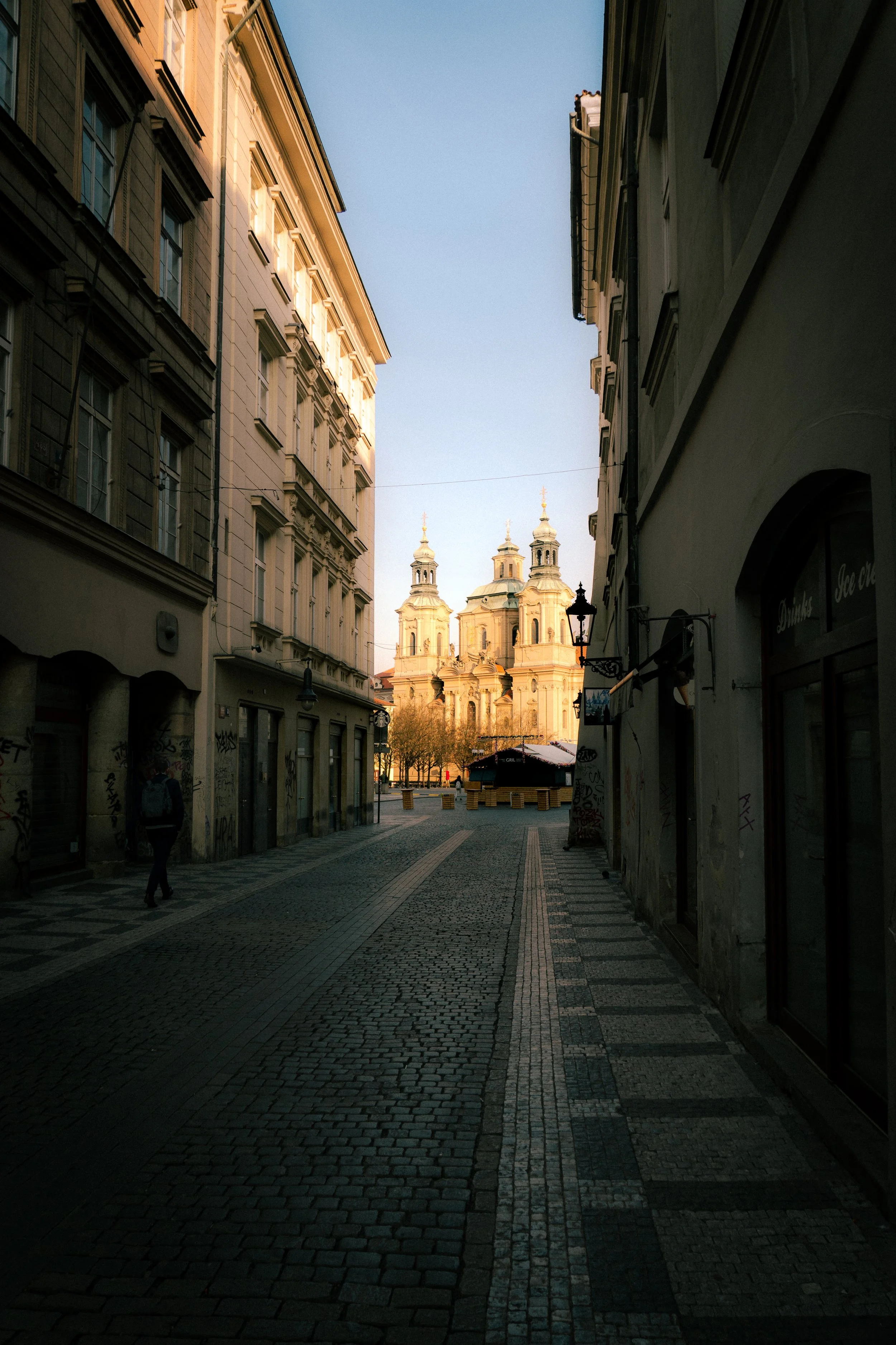 A narrow city street paved with cobblestones, lined with old buildings on either side, and a church with domes and towers visible in the background under a clear sky.