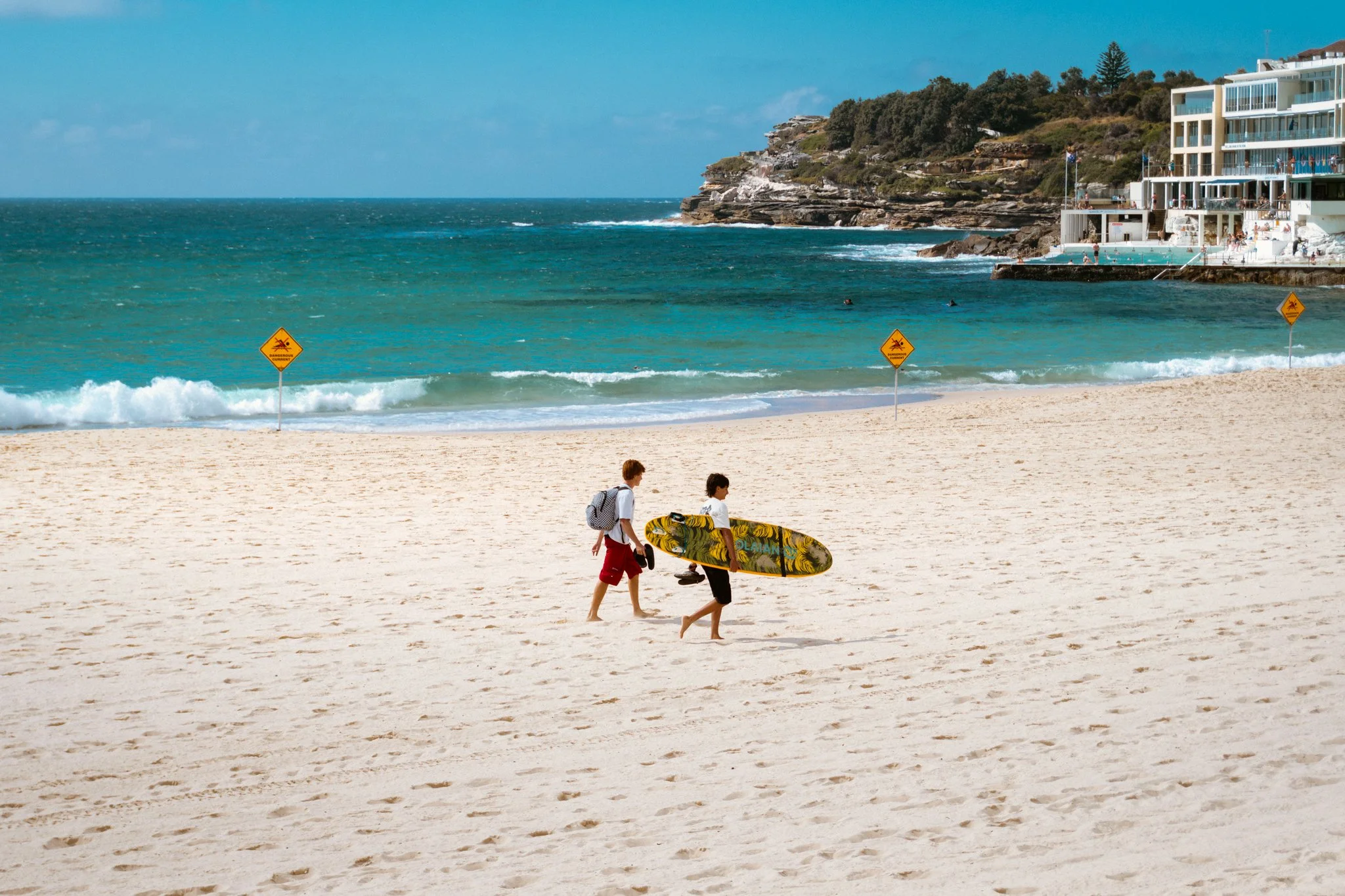 People walking on a sandy beach near the ocean, carrying surfboards, with cliffs and seaside buildings in the background, under a blue sky.