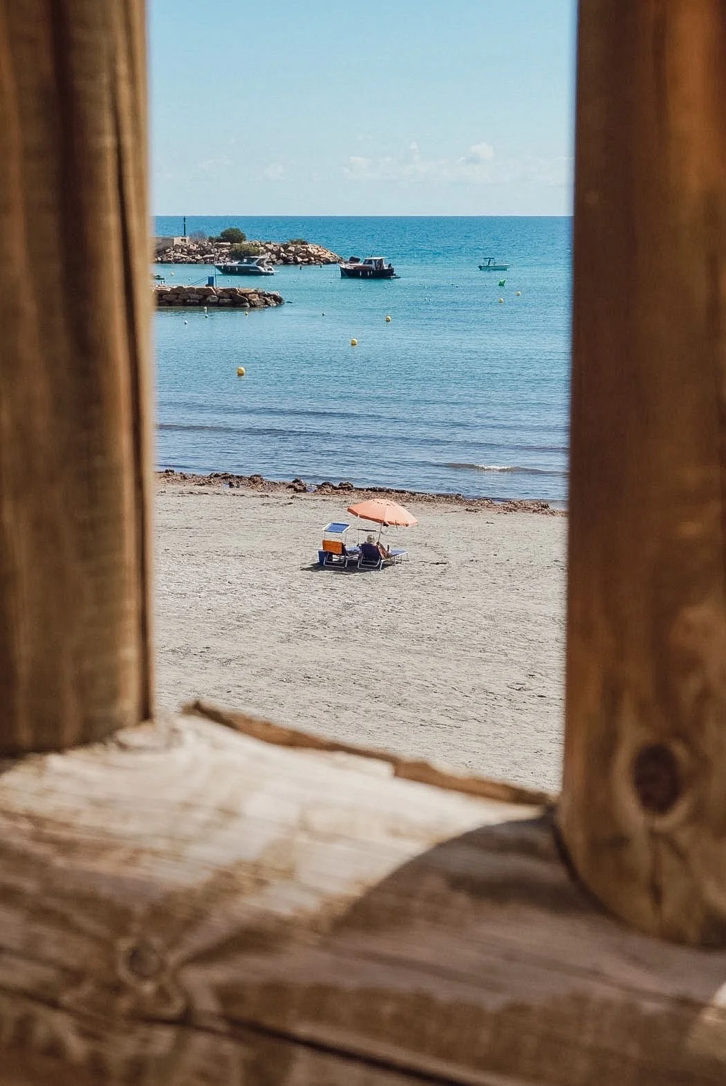 View of a beach scene through a wooden window frame, showing a sandy beach with an orange umbrella and two lounge chairs, calm water with boats, a pier, and rocks in the distance.