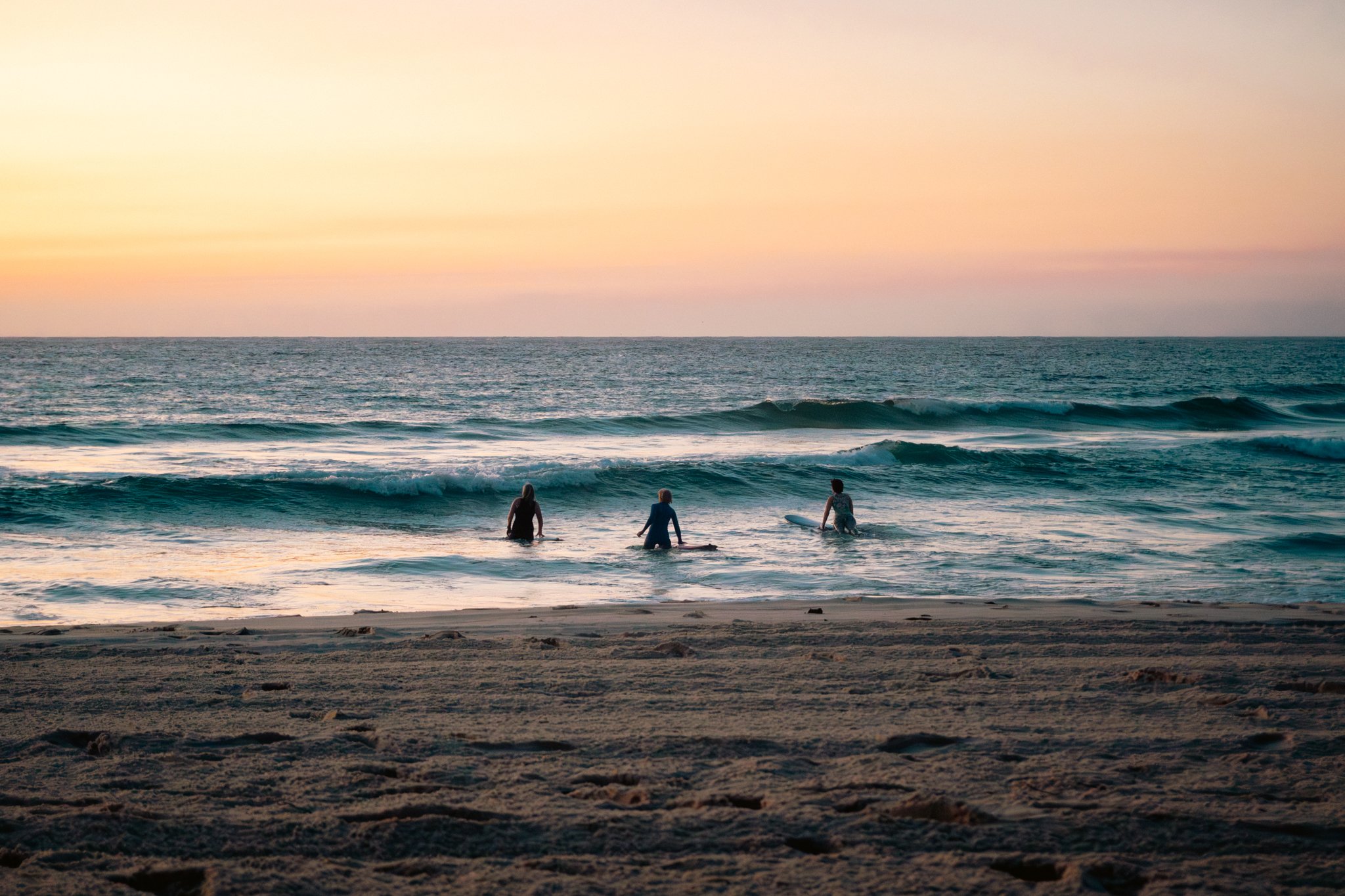 People at the beach during sunset, some with surfboards, facing the ocean and waves.