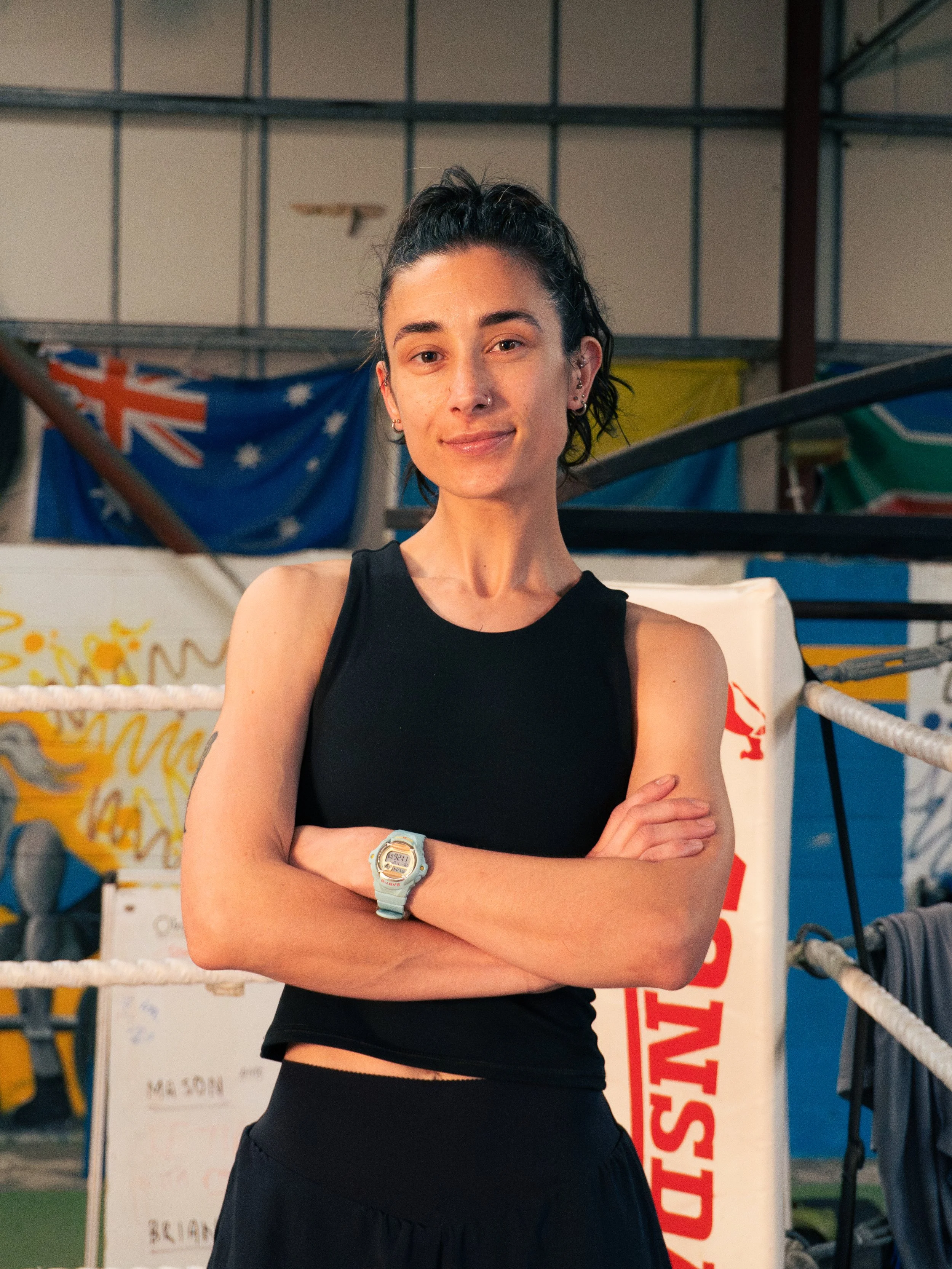 A young woman with dark hair in a high ponytail, wearing a black tank top and black shorts, standing in a boxing gym with her arms crossed and smiling. The background features a boxing ring, Australian flags, and various gym equipment.