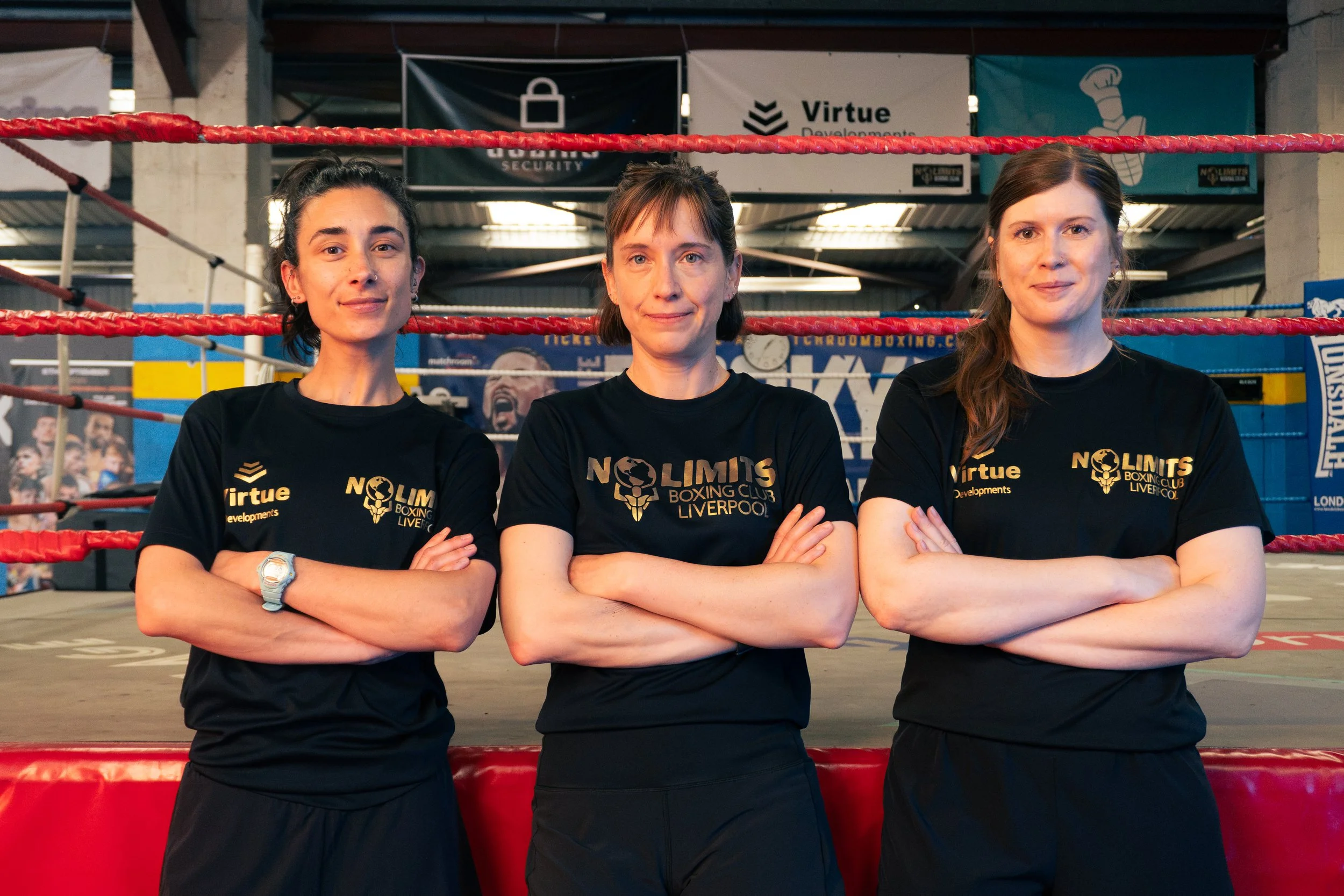 Three women standing in a boxing gym with their arms crossed, wearing black T-shirts with the logo 'NOLIMITS Boxing Club Liverpool' and the word 'Virtue' printed on them. Behind them are boxing rings, banners, and gym equipment.