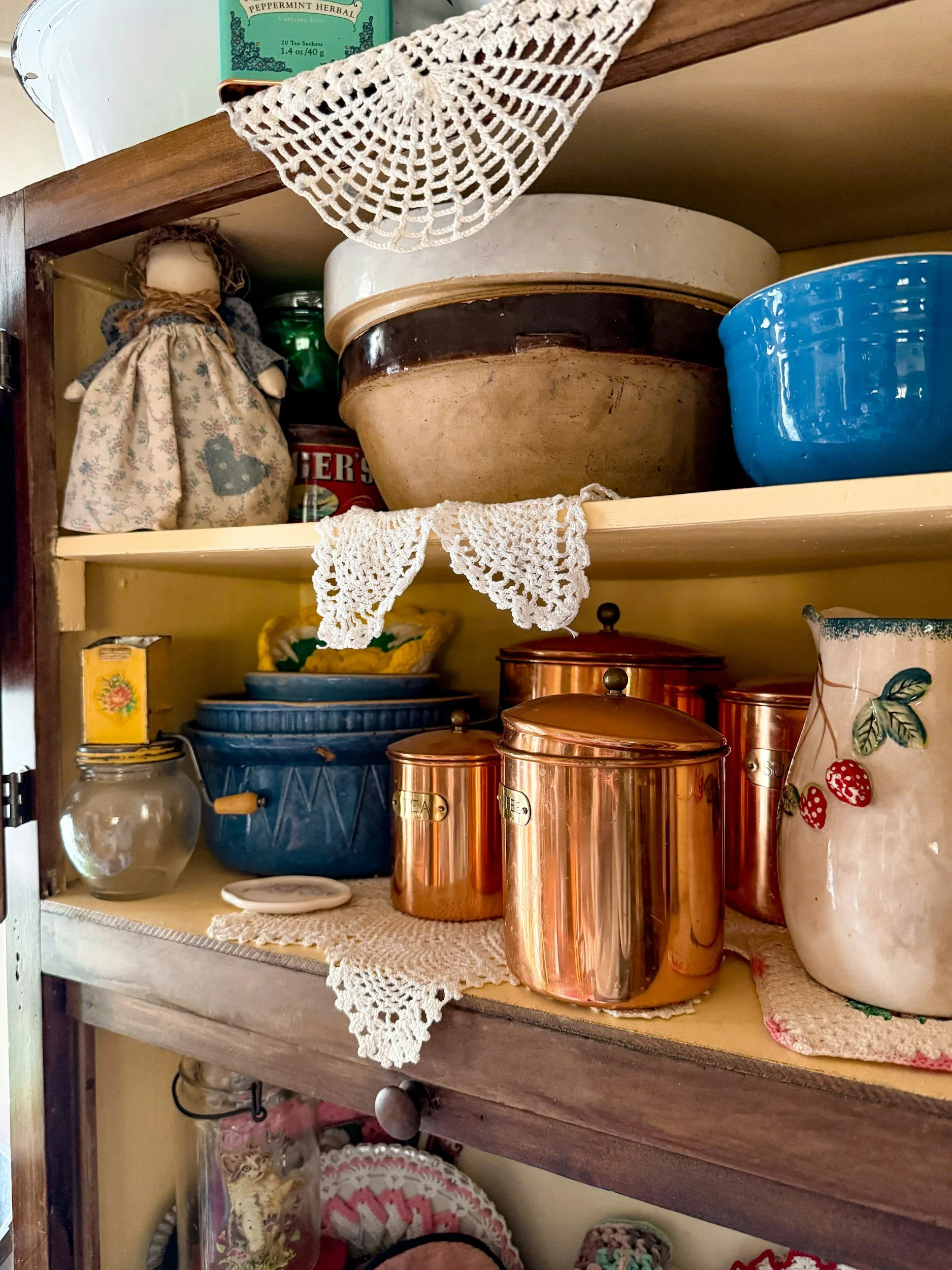 Estantes de madera con utensilios de cocina y objetos decorativos, incluyendo tazas de metal de color cobre, un frasco de vidrio, una taza de cerámica con diseño de fresas, y utensilios en cajas y bolsas. Hay también manteles de encaje y decoraciones de tela.