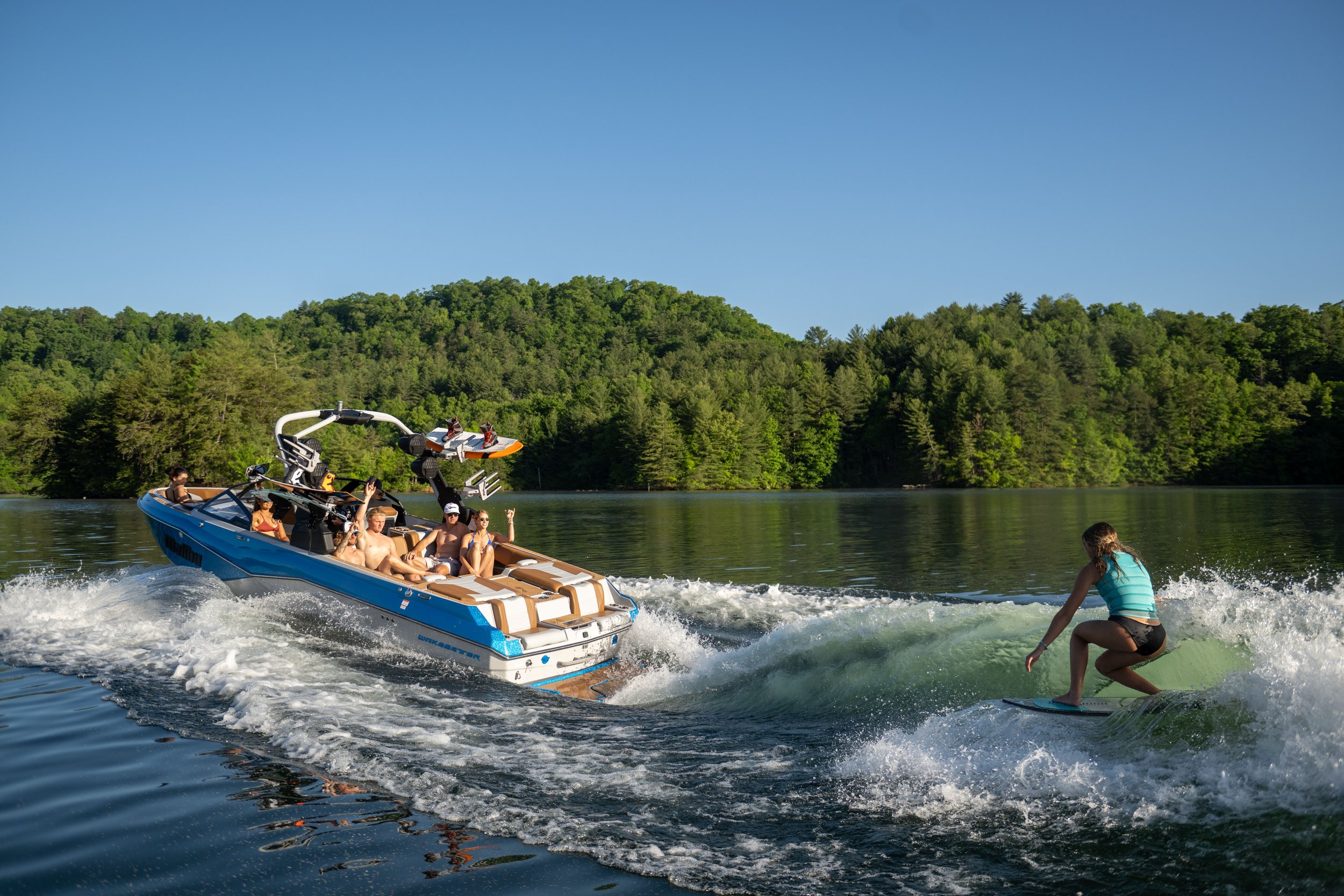 A group of people on a blue and white motorboat enjoying a day on the water, with a person wakeboarding in front of the boat on a lake surrounded by green trees and a clear blue sky.