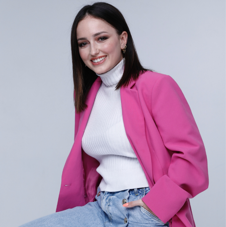 A young woman with shoulder-length dark hair, smiling, wearing a white turtleneck sweater, a bright pink blazer, and light blue jeans, standing against a plain light gray background.