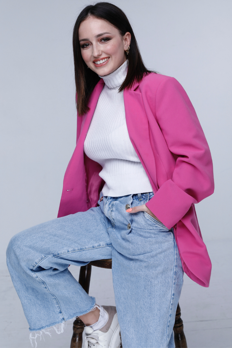 A young woman with dark hair wearing a pink blazer, white turtleneck sweater, light blue ripped jeans, and white sneakers, sitting on a wooden chair against a plain light gray background, smiling at the camera.