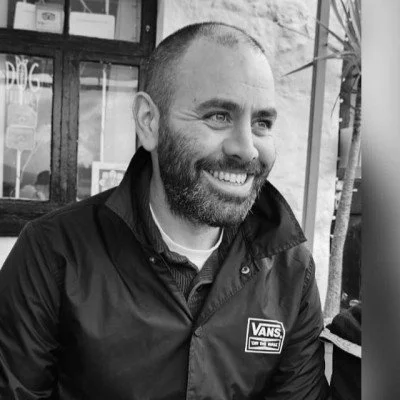 A smiling man with a beard and short hair, wearing a jacket with a Vans logo, outside a building with large windows.