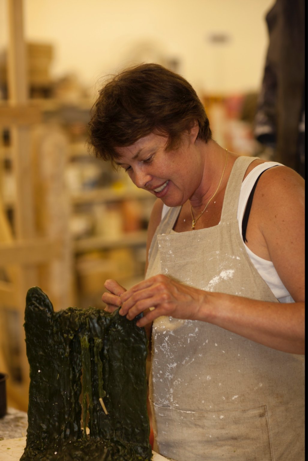 A woman in a beige apron smiles while working with a large sculpture made of modelling wax.
