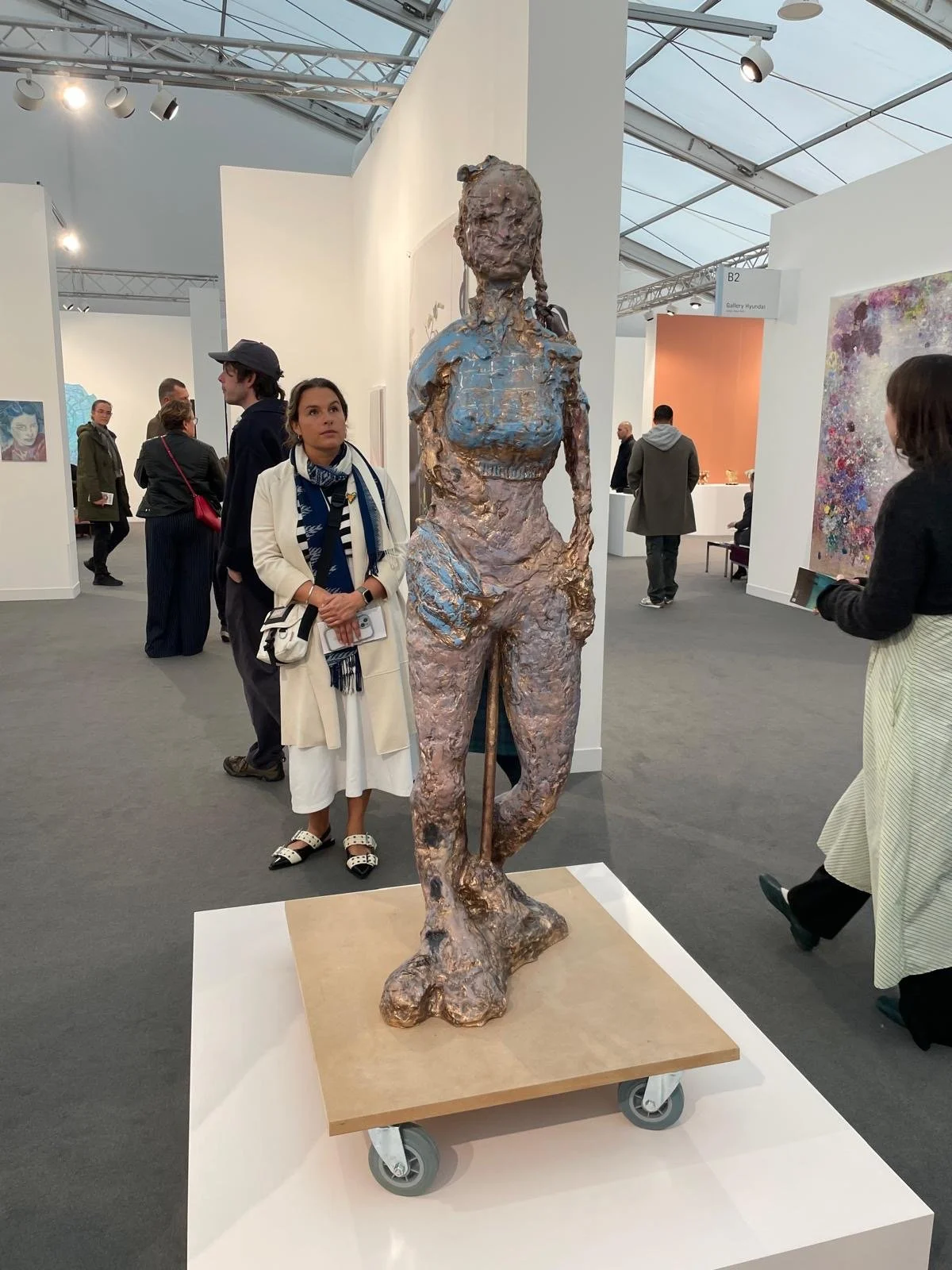 Voyager b by Rebecca Warren. A bronze sculpture of a woman with an exaggerated, rough texture, painted in shades of gold and blue, standing on a wheeled platform at Frieze Art Fair 2025.