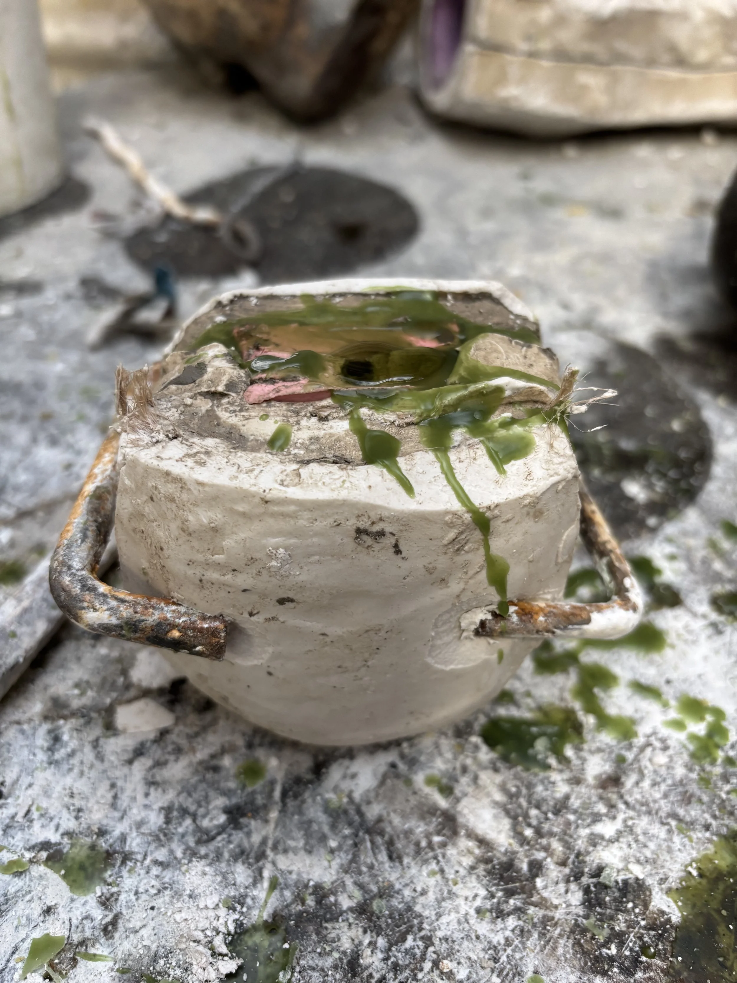 A small plaster mould containing liquid green painting wax positioned on a dirty, dusty surface outdoors, with assorted objects in the background.