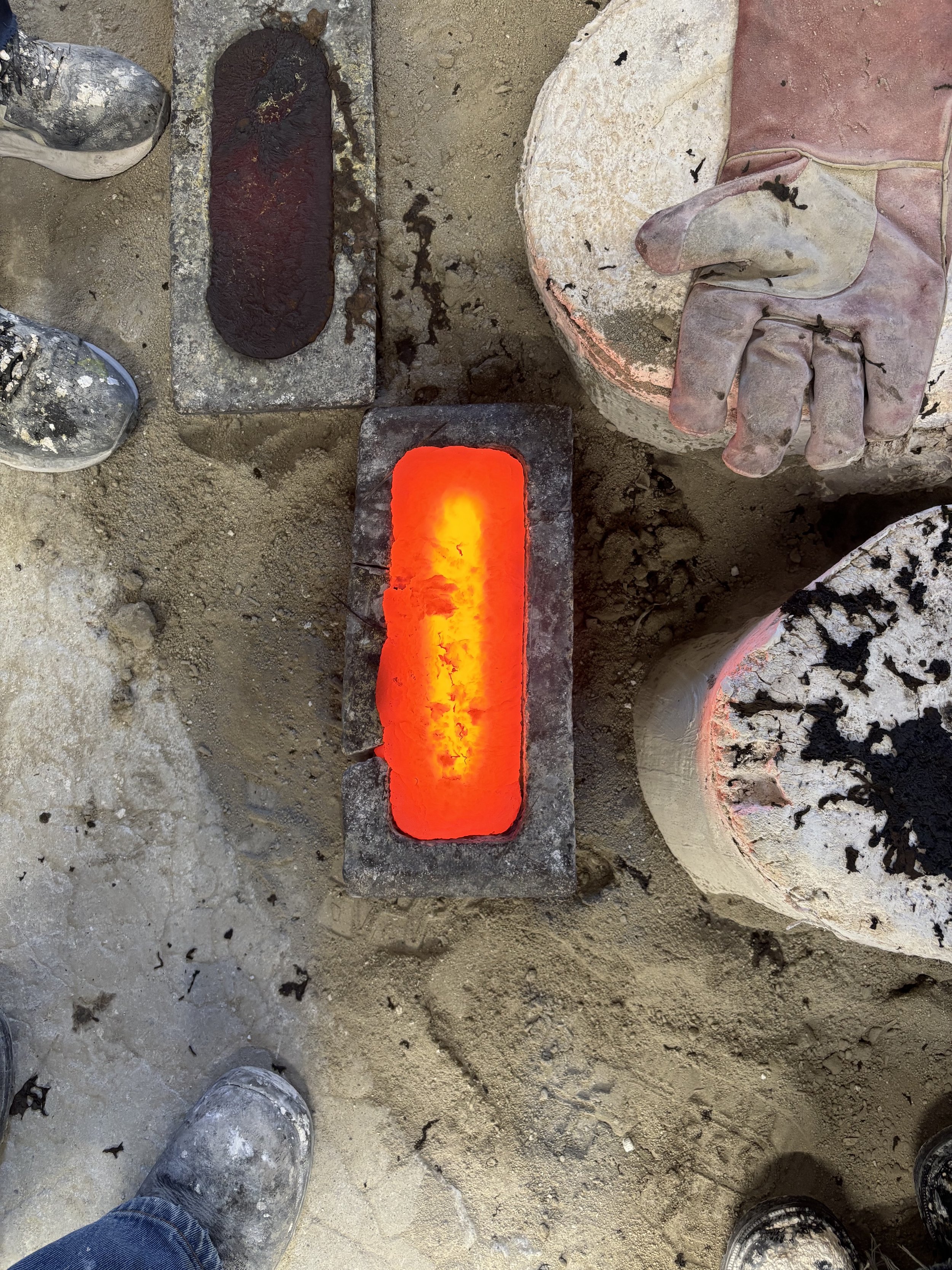 Close-up of a recently poured ingot glowing red, surrounded by various moulds, with two pairs of dirty work shoes visible at the bottom.