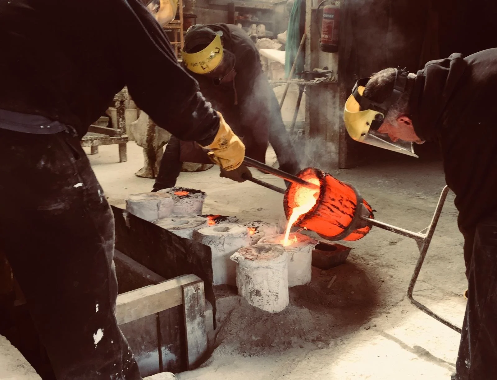 Three foundry workers wearing protective gear including masks and gloves, pouring molten metal from a crucible into a mold in a workshop with metal tools.