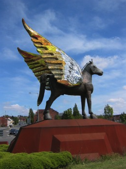 Pegasus by Andrew Logan. A large bronze sculpture of a winged horse with colourful wings on a red base in an outdoor setting with a cloudy sky.