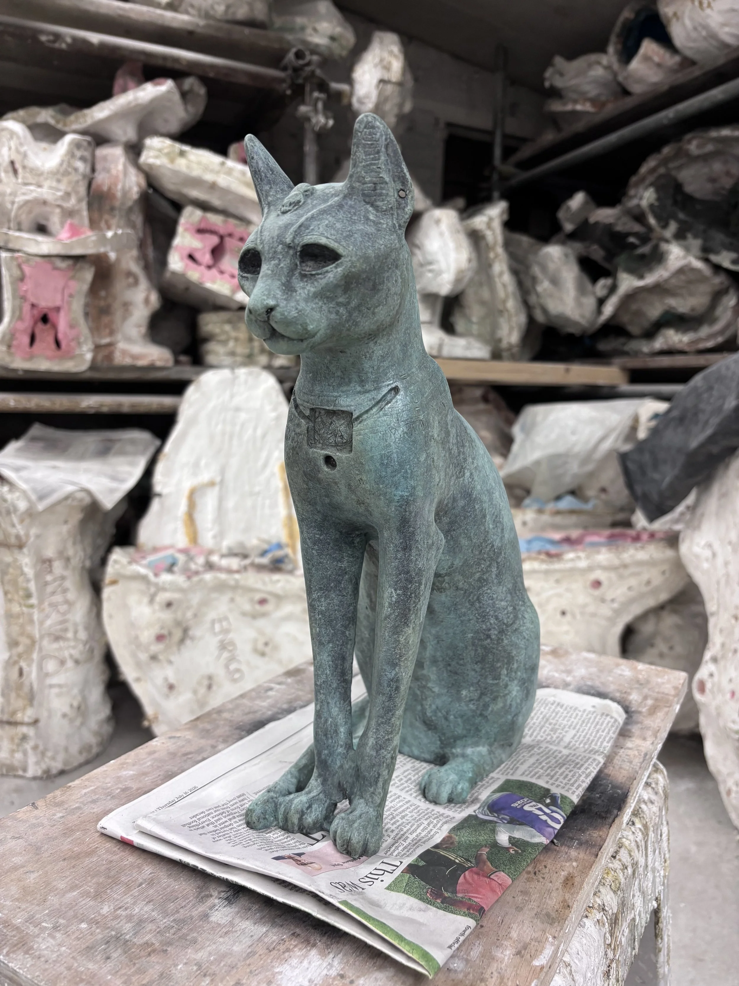 Gayer Anderson Cat. A bronze statue of a cat sitting on a wooden surface covered with newspapers, in a workshop with clay and plaster molds in the background.