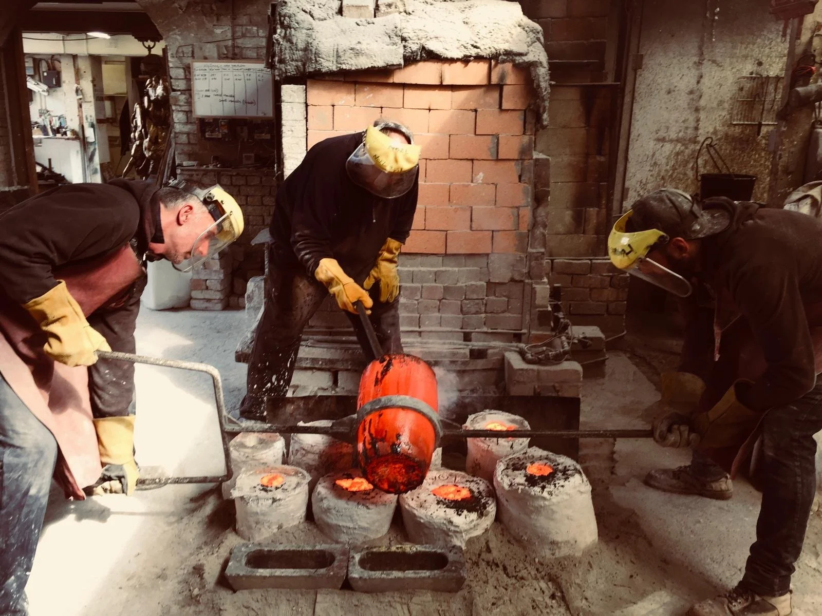 Three foundry workers wearing protective gear including masks and gloves. Two are pouring molten bronze metal from crucible into moulds made of plaster. Part of the lost wax bronze casting process. Inside a casting foundry.