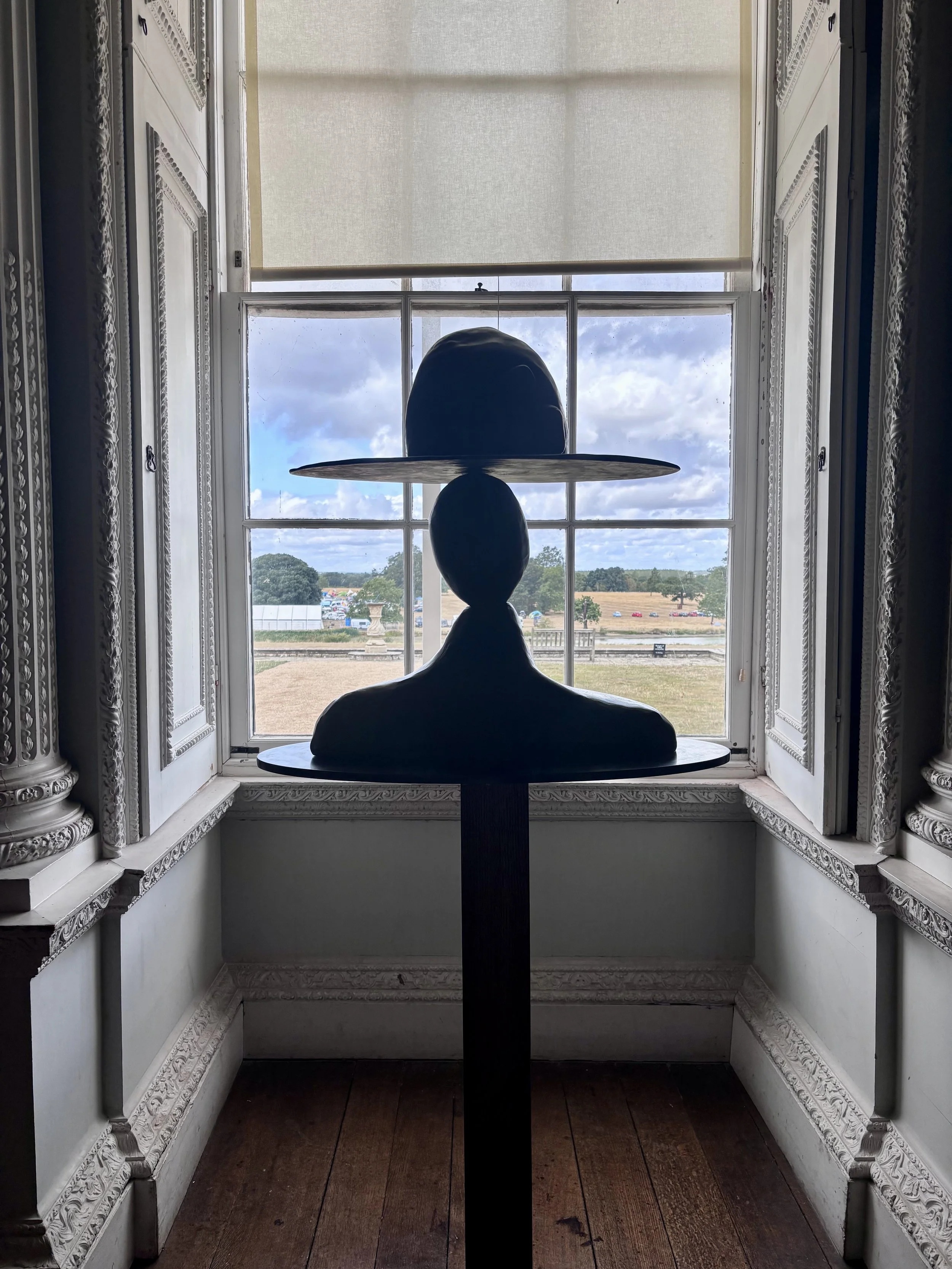 You are her by Enrico David. An interior view of a window with carved wood detailing and a statue of a person wearing a hat, with the sky and landscape visible outside.